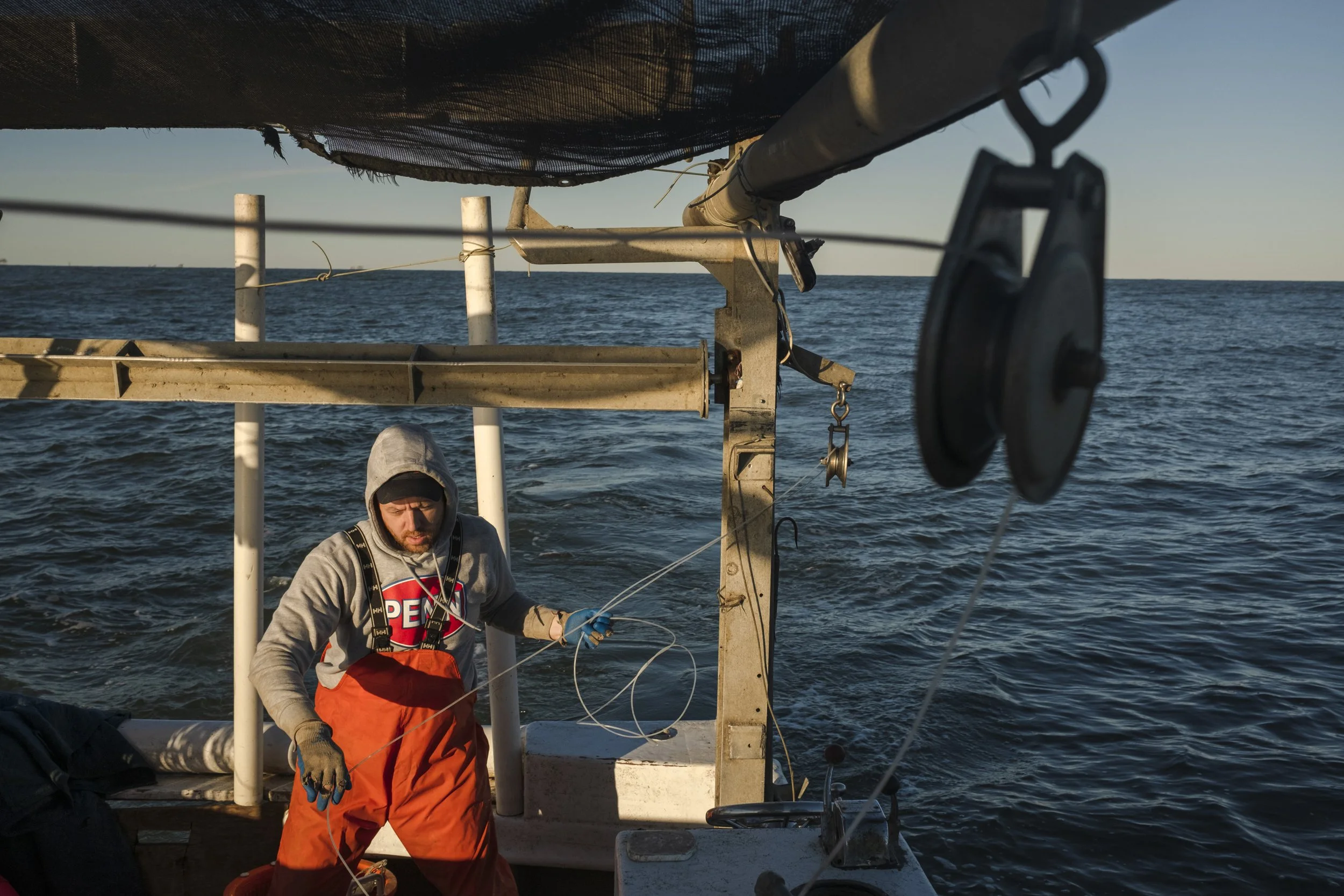 A person in orange overalls and a gray hoodie on a boat, handling fishing gear or equipment, with the ocean and a clear sky in the background.