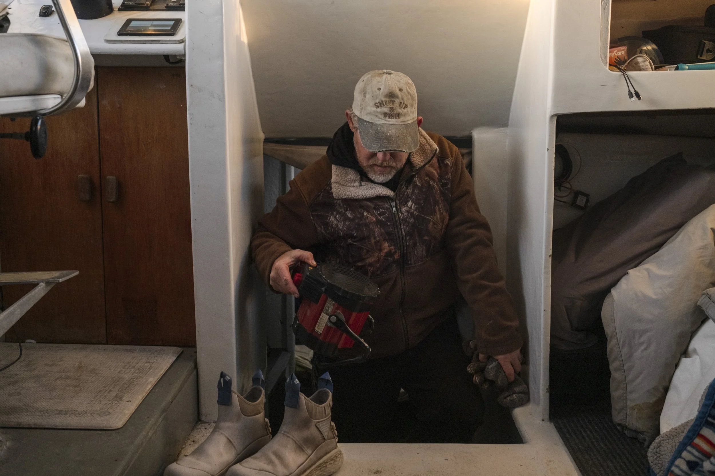 Man wearing a cap, jacket, and glasses kneeling in a small space, holding a red and black spotlight, with work boots in front of him, surrounded by cluttered shelves and household items.