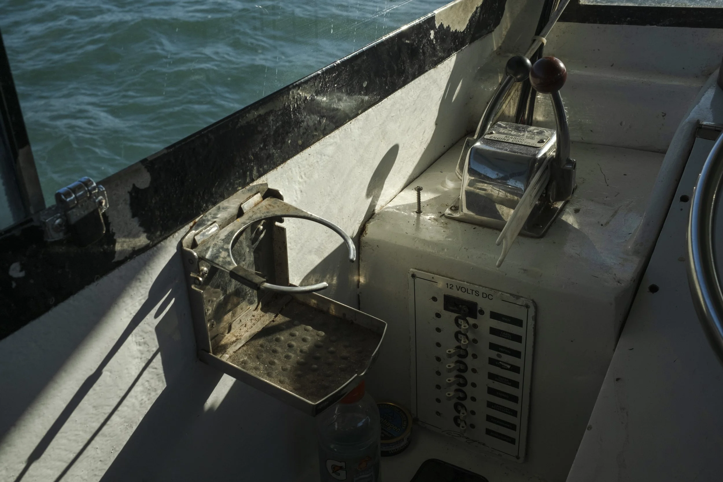 Close-up view of a boat's control panel with a throttle handle, a small metal tray, a water bottle, and an electrical panel labeled '12 VOLTS DC' with switches, next to water with gentle waves visible outside the boat.