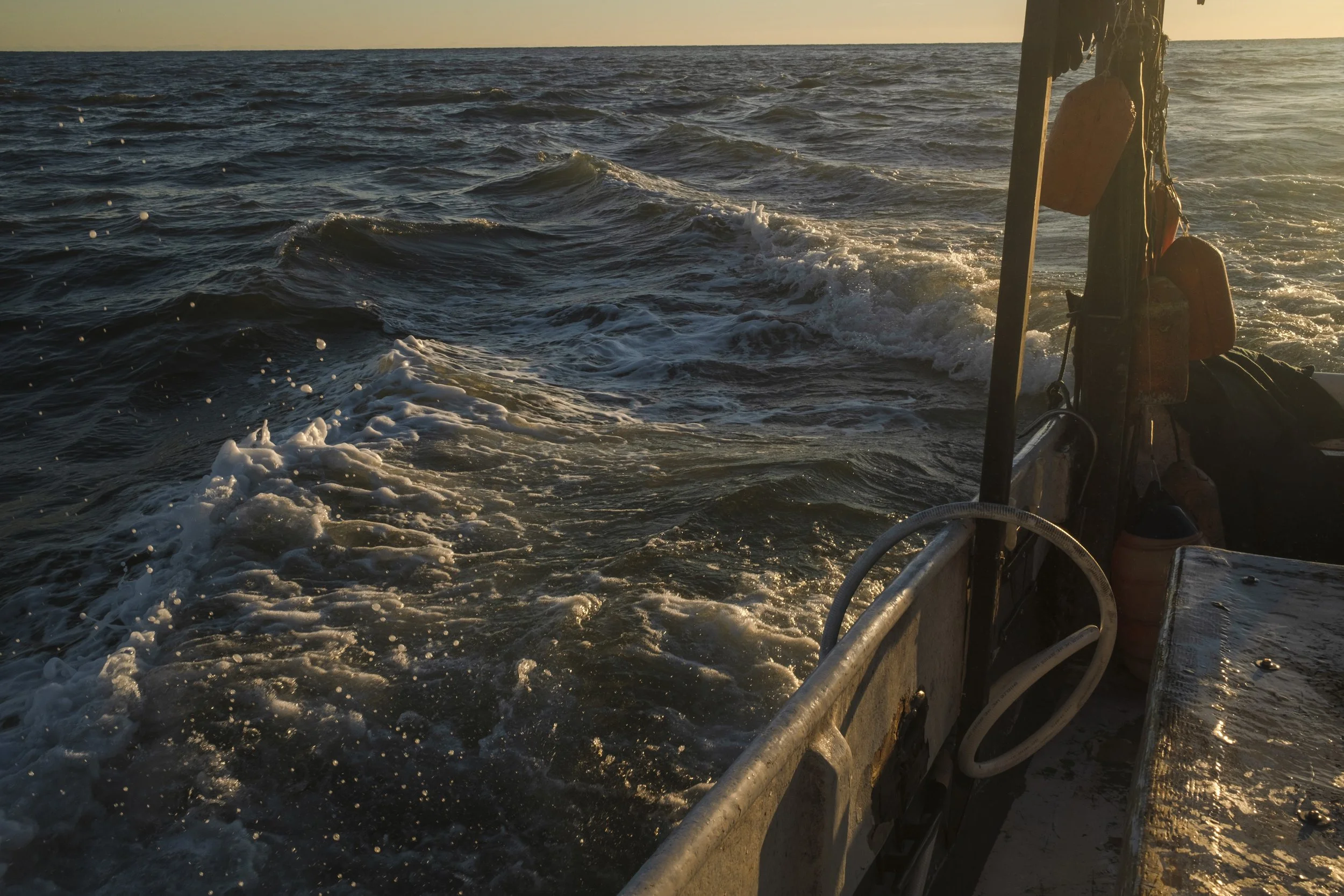The photo shows the view from a boat, capturing the ocean waves at sunset with the boat's rear section, including lifebuoys, ropes, and part of the deck, in the foreground.