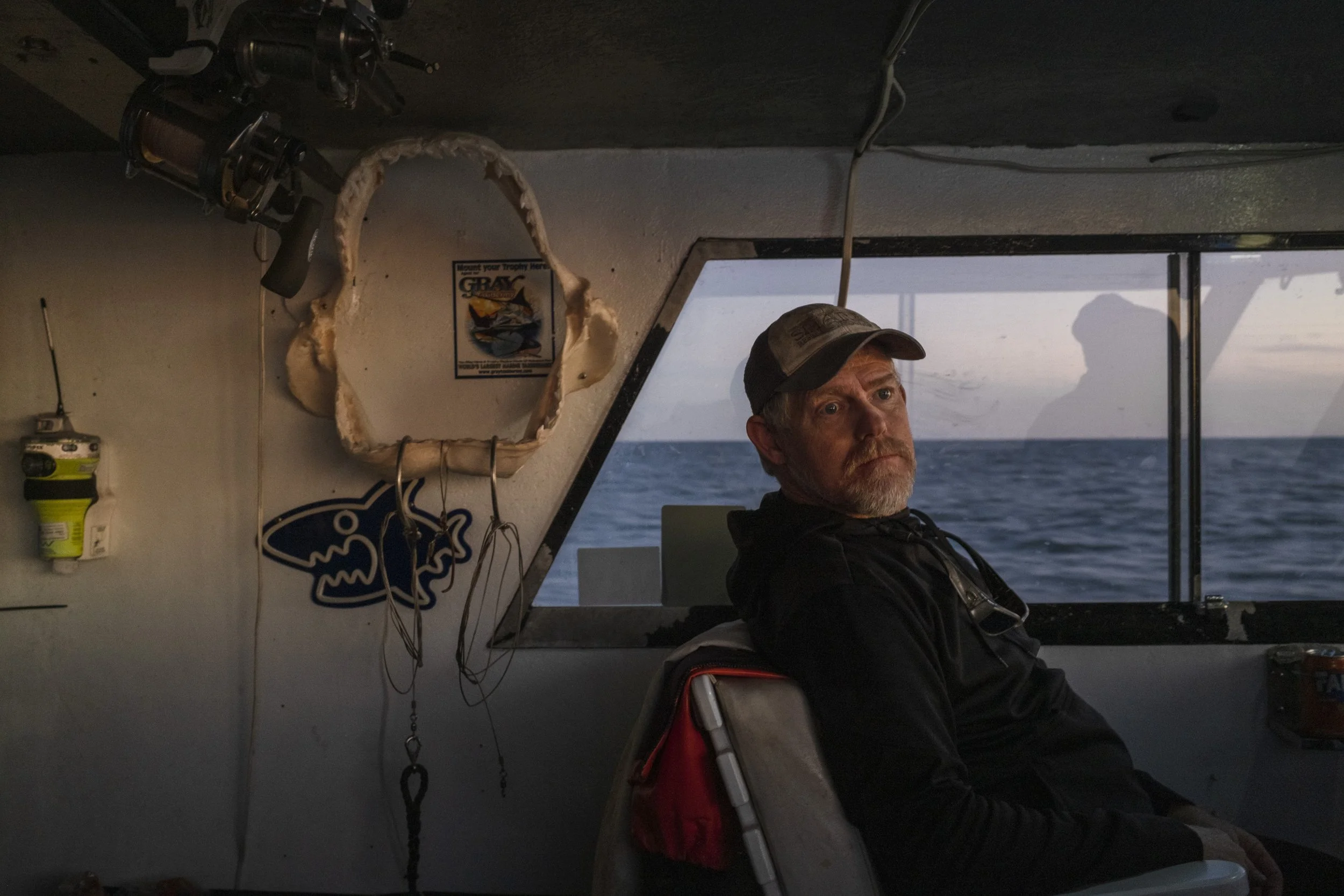 A man with a beard and baseball cap sitting on a boat looking towards the camera, with the ocean and sky in the background. The boat interior has fishing equipment and fishing-related decorations.
