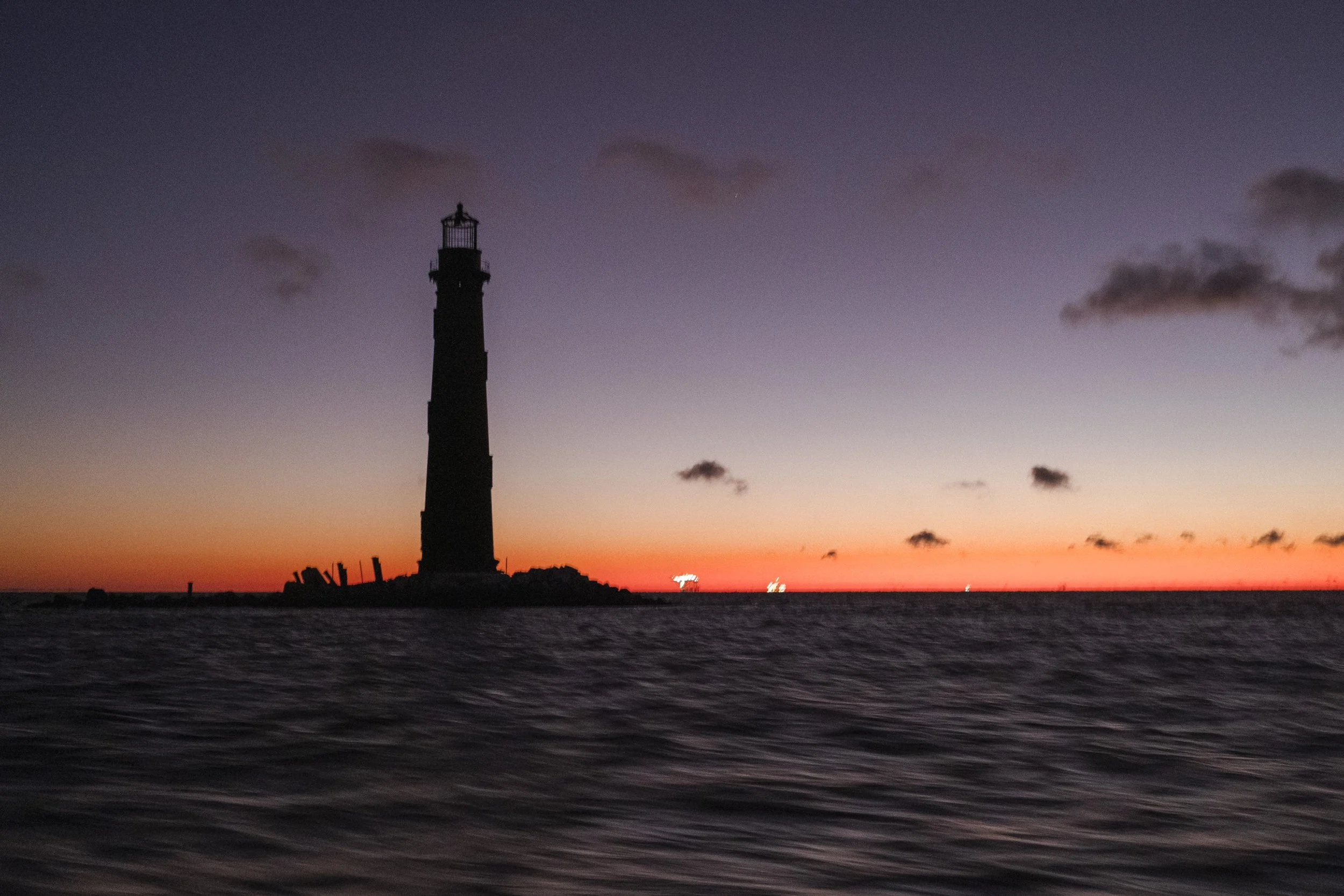 Lighthouse silhouetted against a colorful sunset sky over the ocean with gentle waves in the foreground.