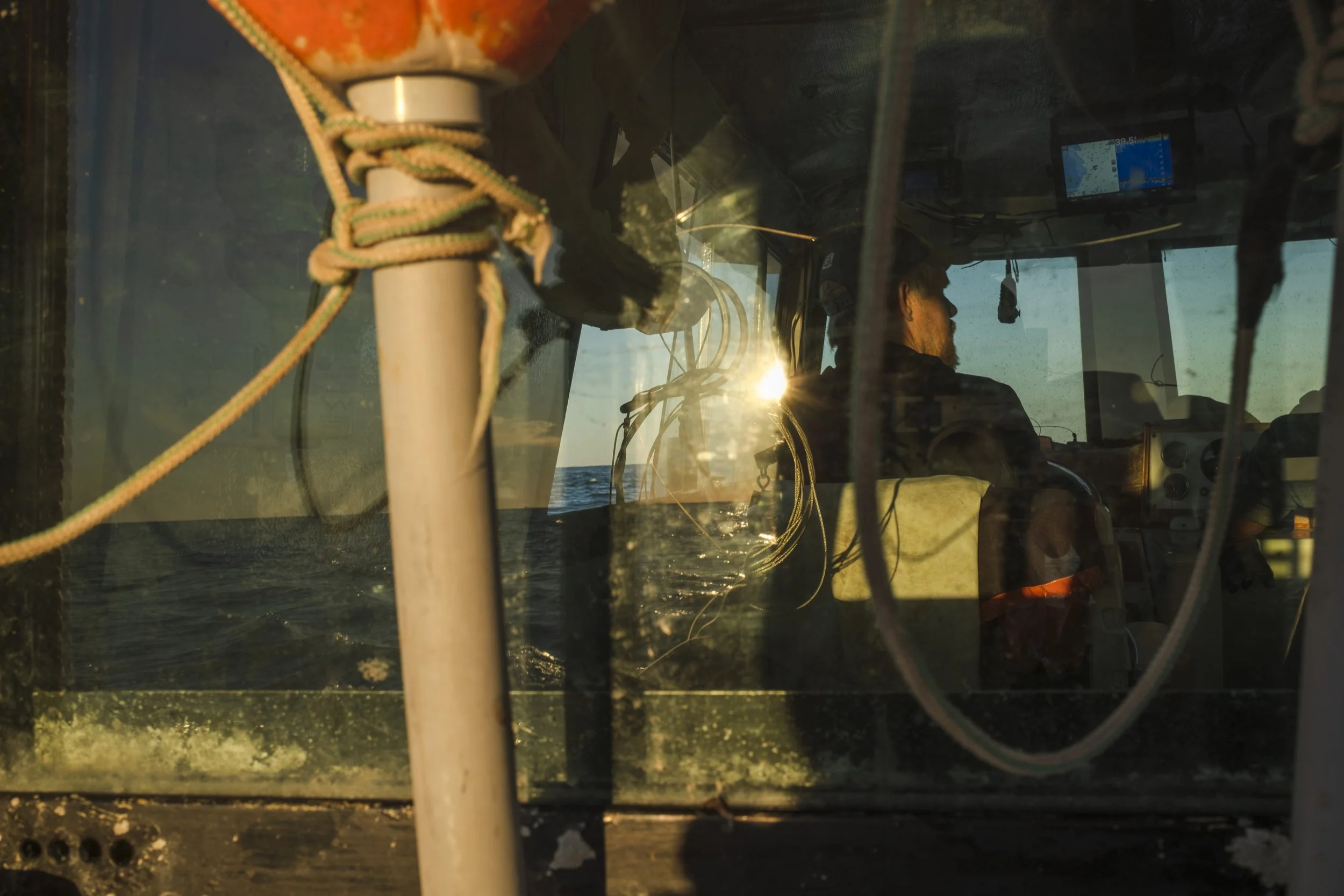 View through the front window of a boat showing the silhouette of a person at the helm with the setting or rising sun reflecting on the sea outside.
