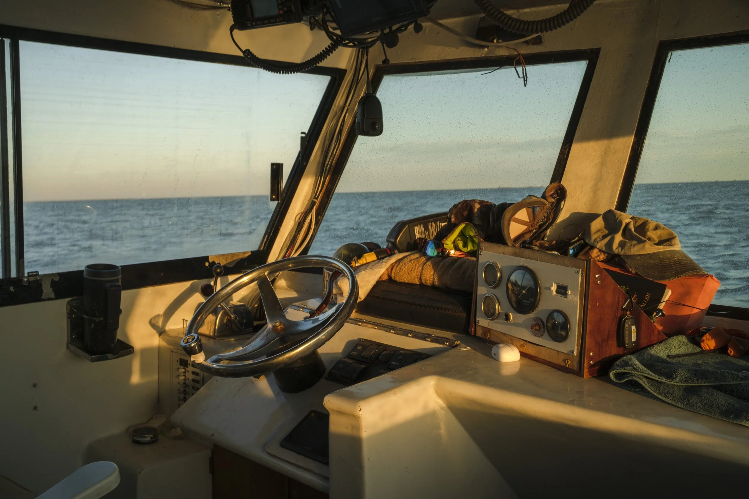 View from the boat's bridge showing the ocean and sky through the windows, with the boat's steering wheel, control panel, and various items like a hat, towel, and gear on the dashboard.