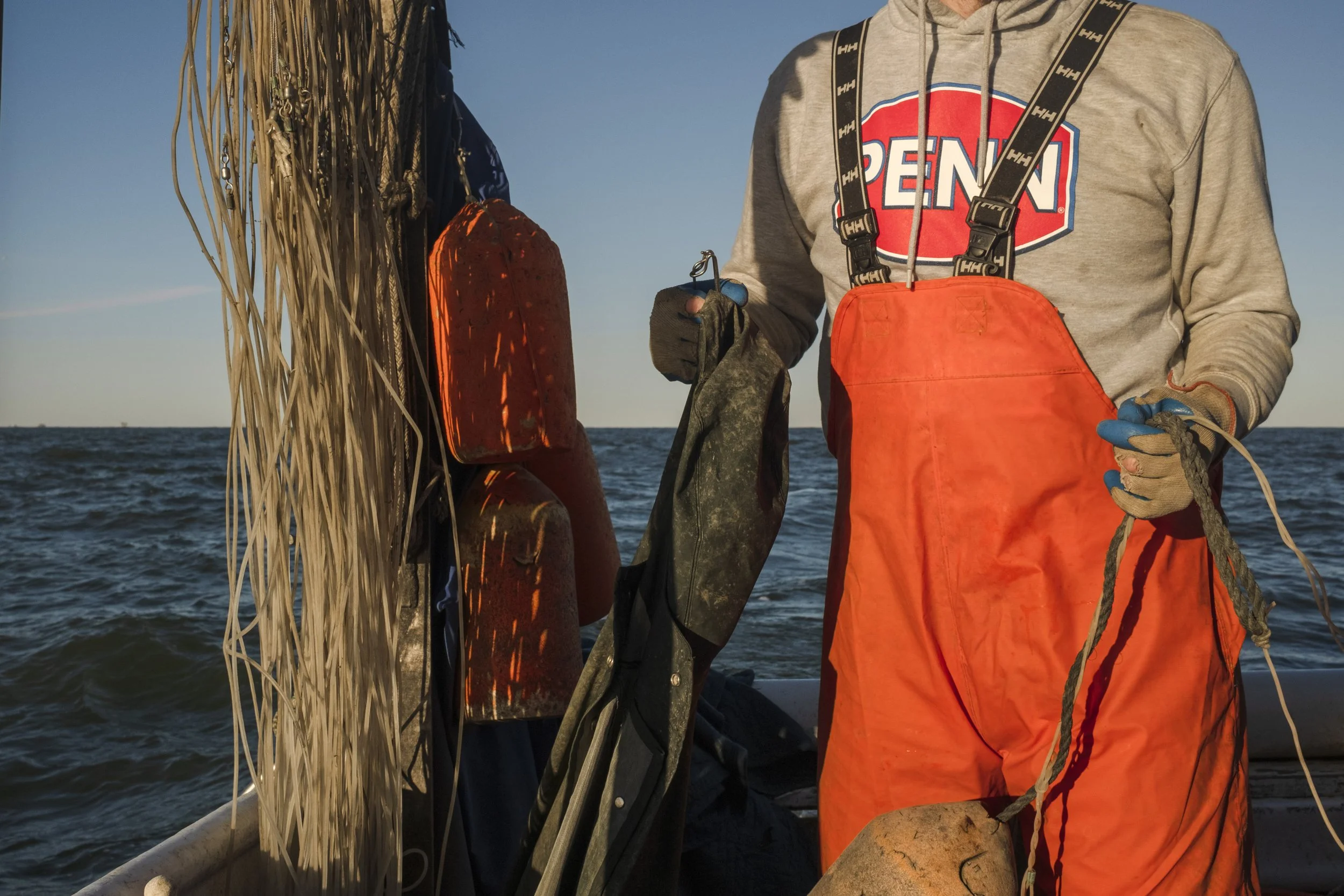 Person on a boat wearing an orange waterproof bib and gray hoodie with a PENN logo, holding a rope, surrounded by life jackets and fishing gear, with the ocean in the background.