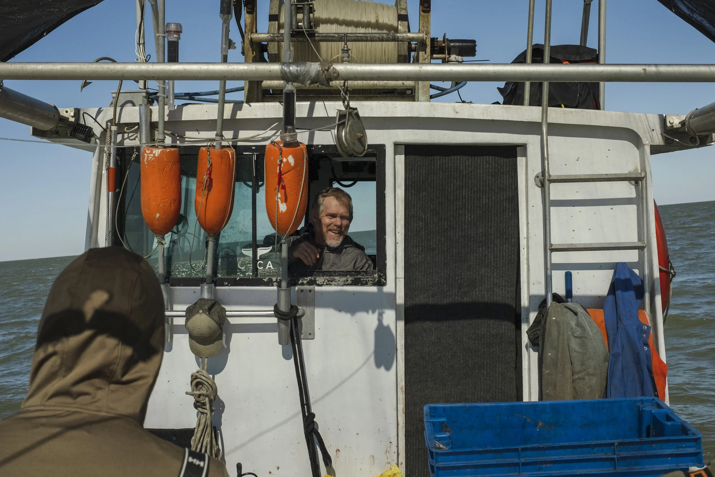 Man smiling in the wheelhouse of a boat, with orange buoys hanging outside, on a body of water under a blue sky.