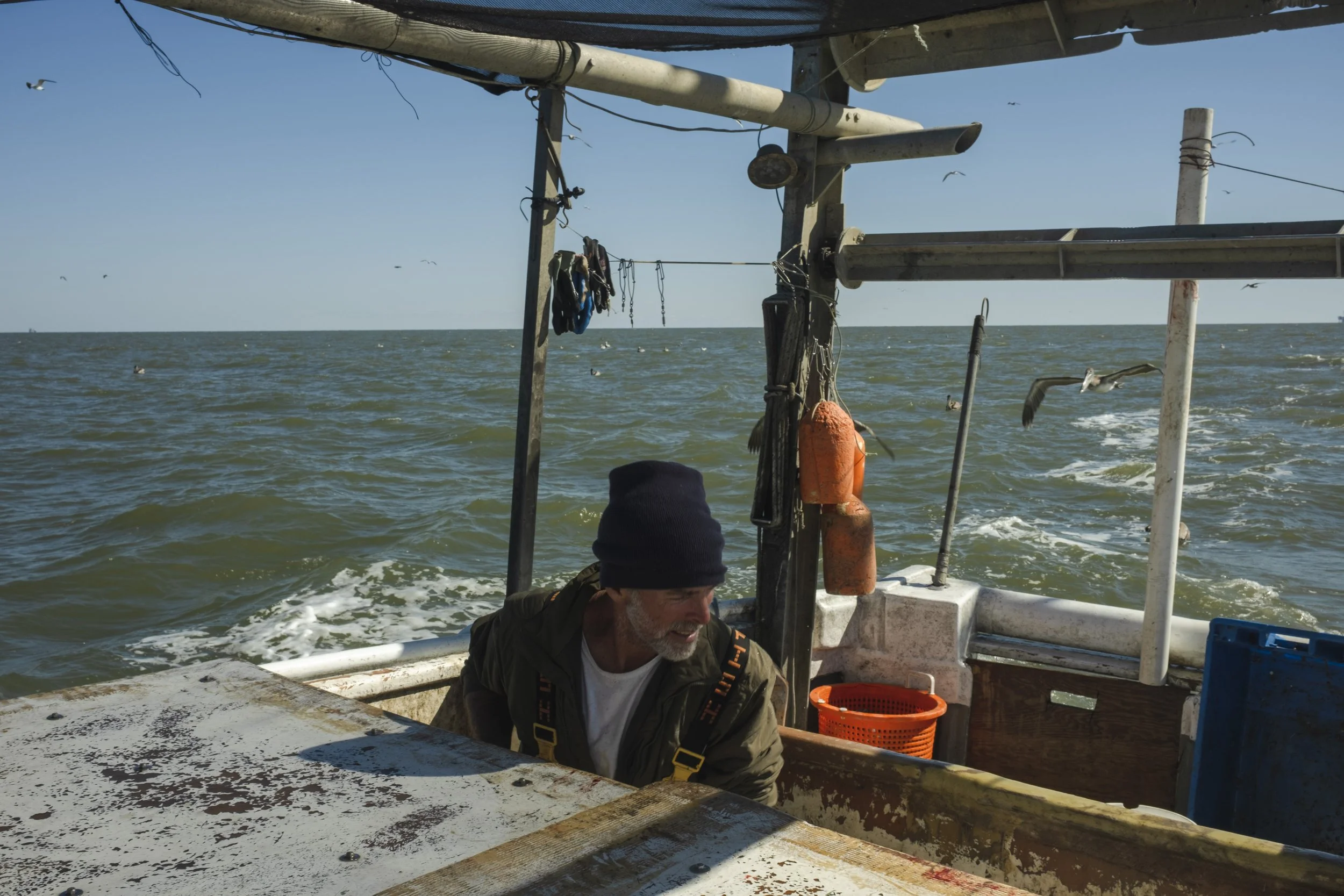 A man wearing a black beanie and jacket on a boat at sea, with birds flying and fishing equipment around him.