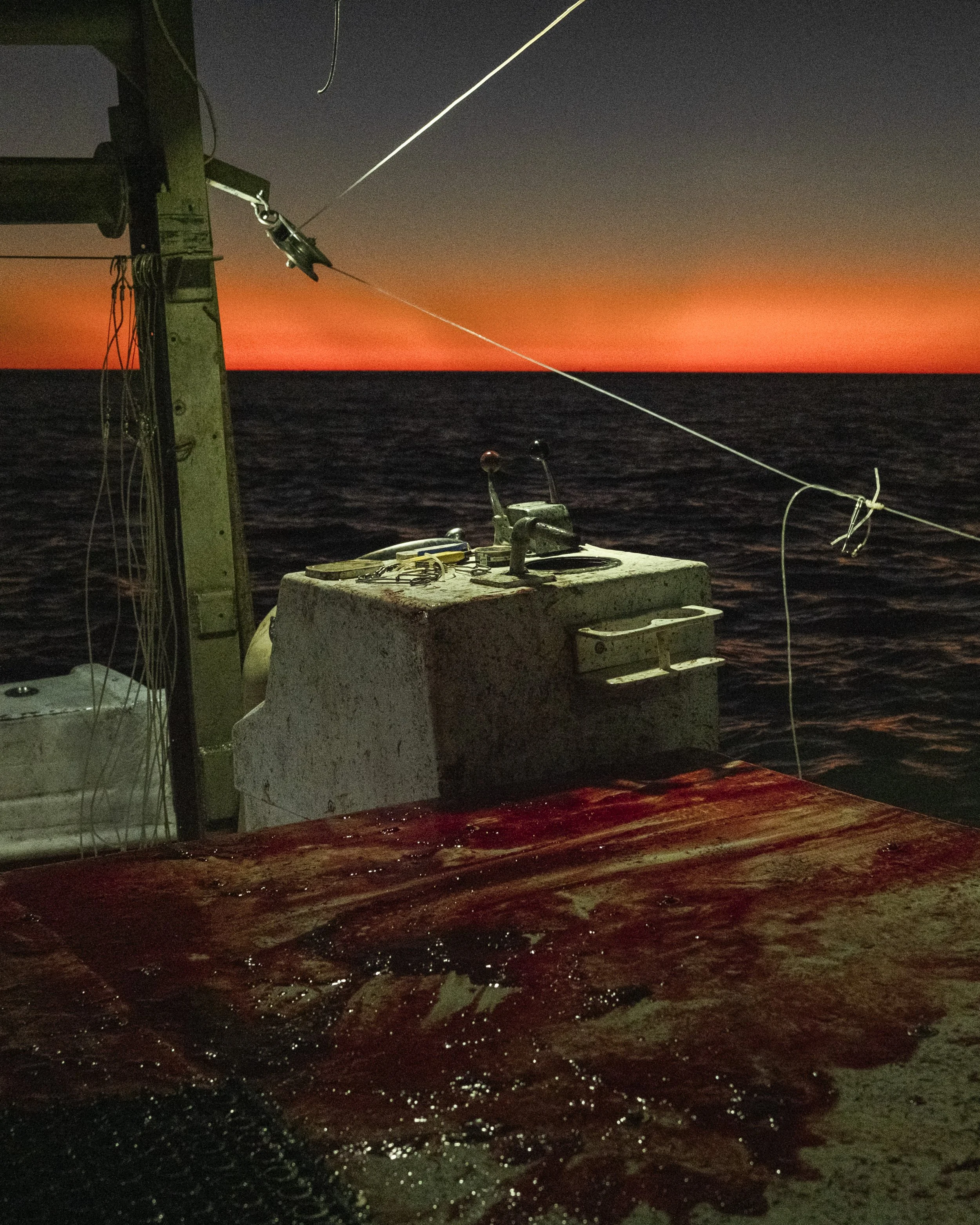 View of the deck on a boat at sunset, with fishing poles rigged, and the ocean in the background under a colorful sky.