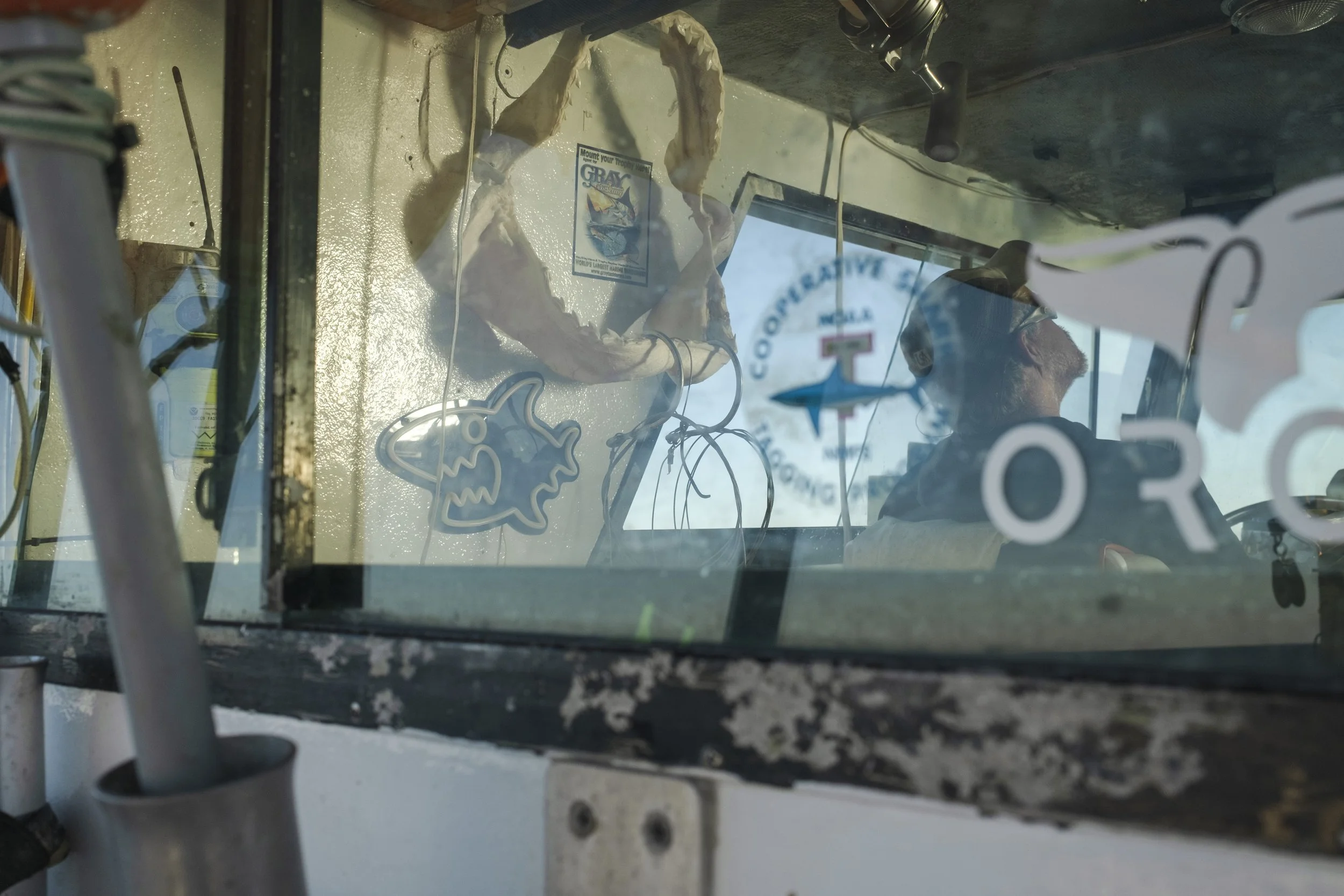 View through a vehicle window showing a person wearing sunglasses and a cap, with a logo behind them, and a decorative hanging shark shape on the wall.