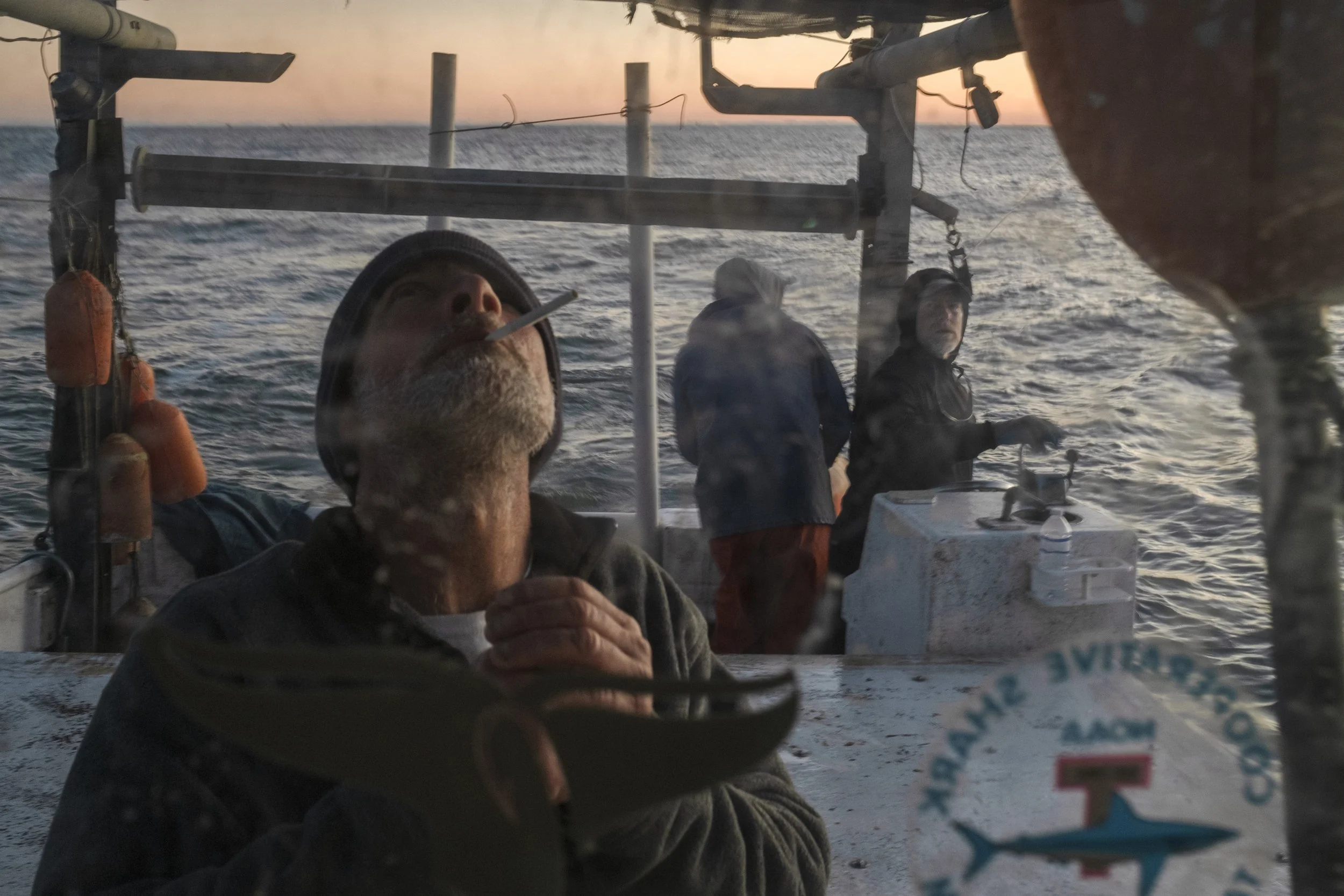 A man on a boat smoking a cigarette and holding a bowl, with other people in the background looking at the ocean during sunset.