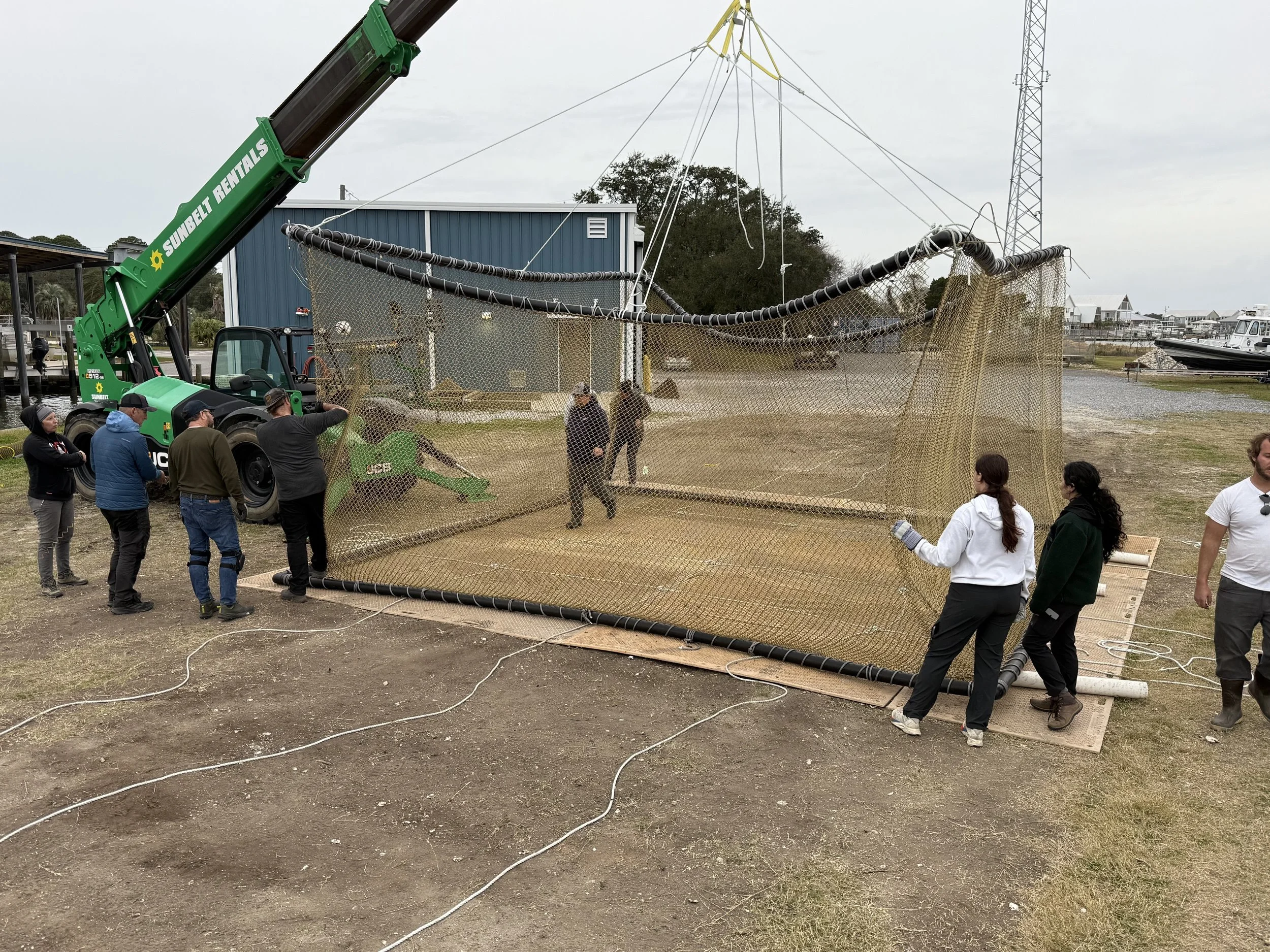 People working on installing or maintaining a large fishing net with a crane at a marina or waterfront area.