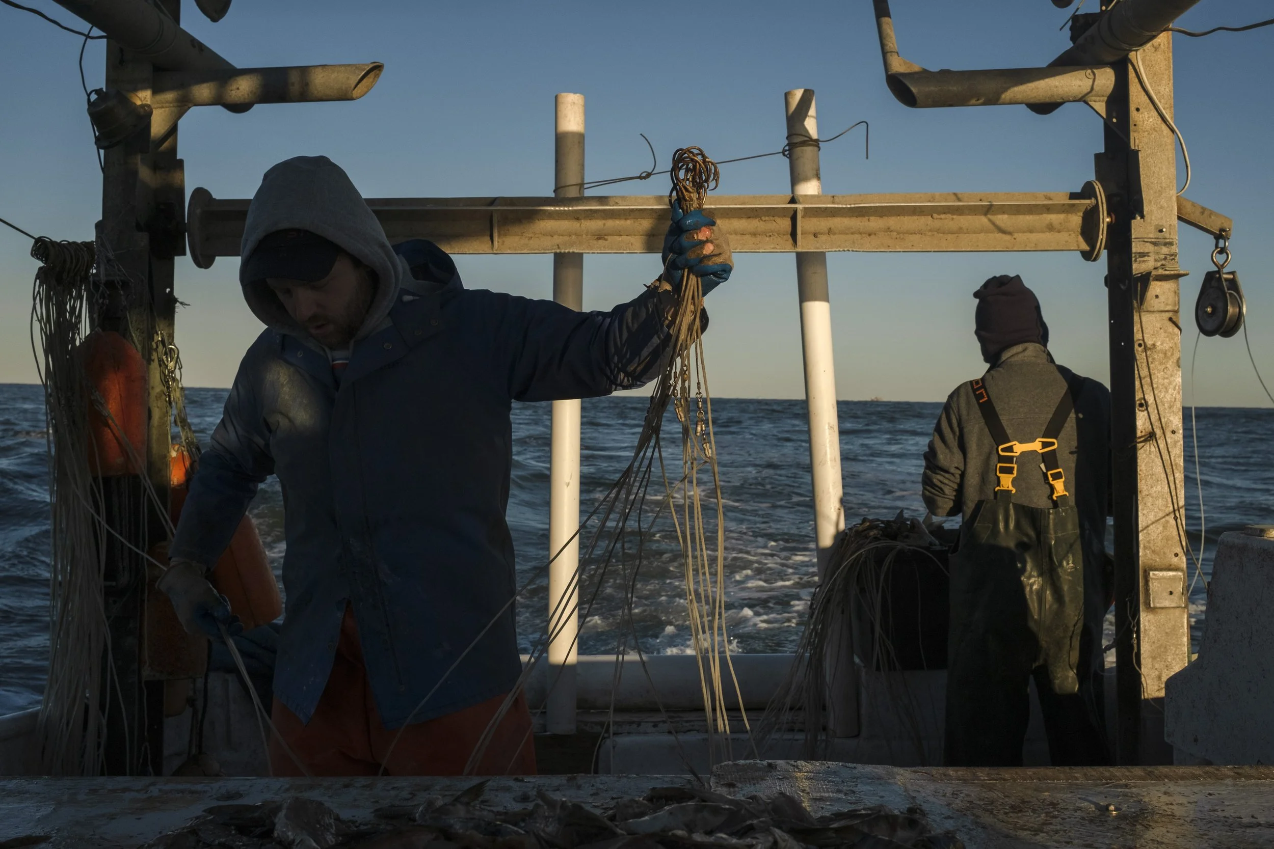 Two fishermen on a boat at sea, one handling fishing nets and the other looking away, during sunset.