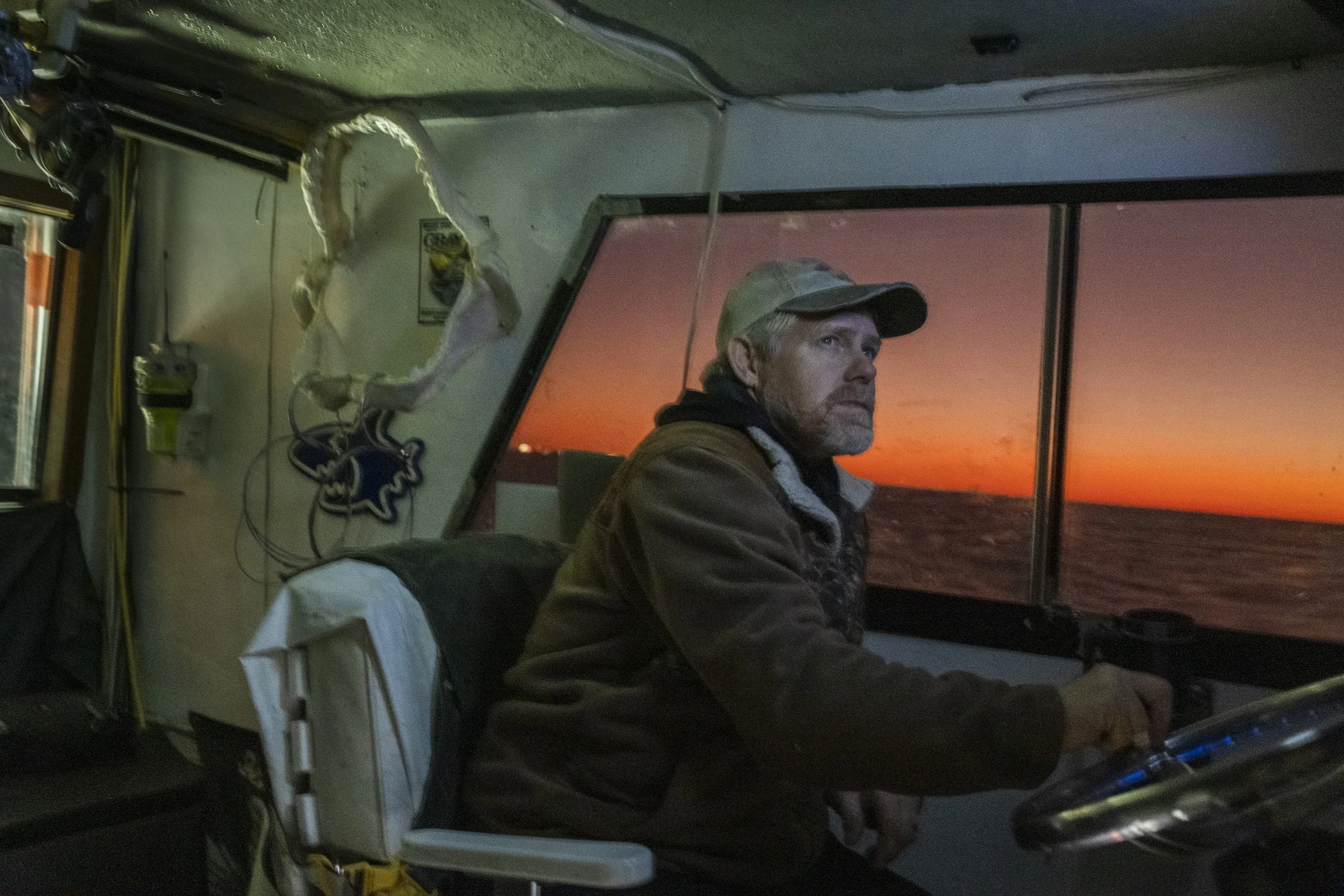A man with a beige cap and brown jacket sitting inside a boat's cabin, looking thoughtfully through the window at a sunset over the ocean.