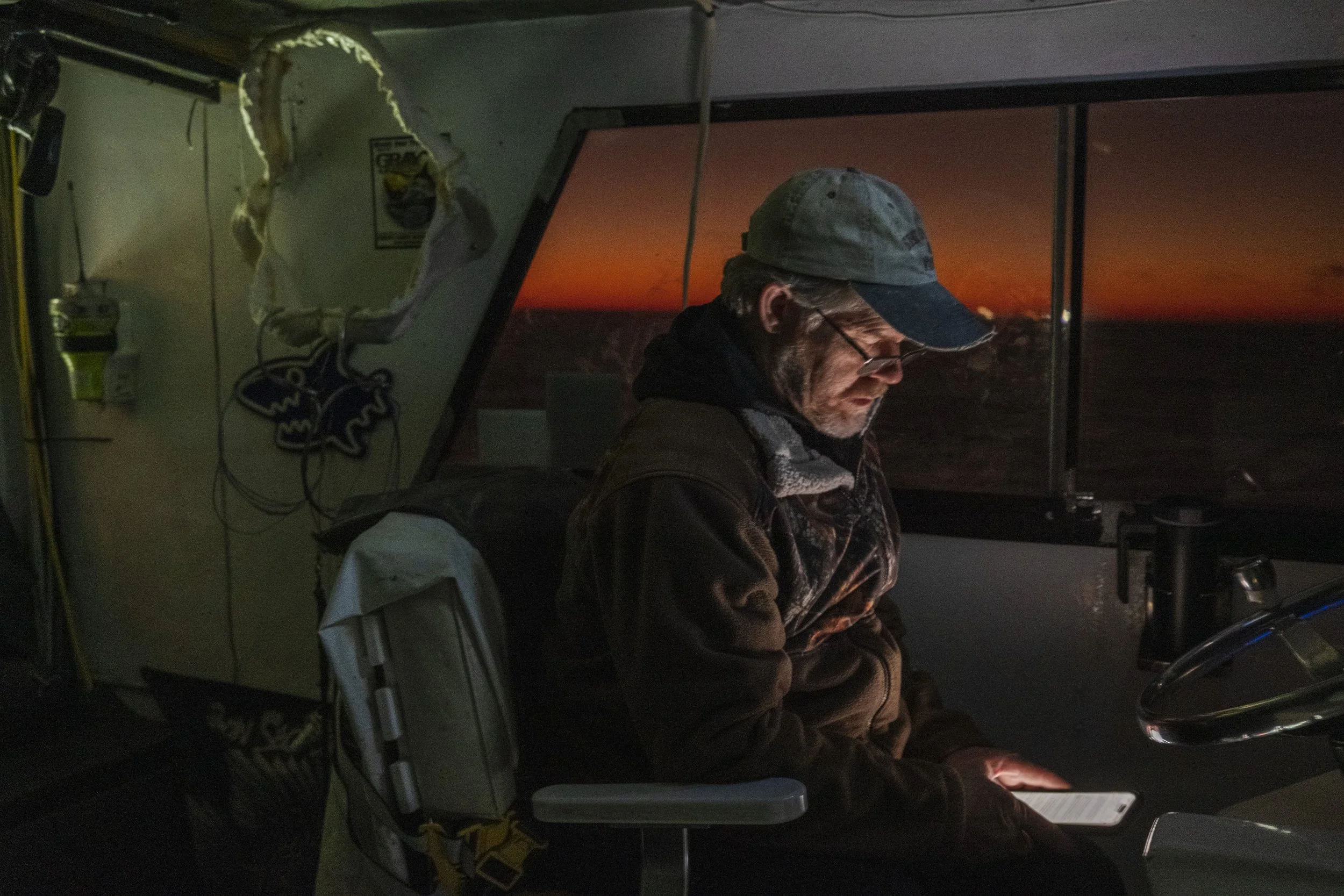 An older man sitting in a vehicle at sunset, looking at a smartphone with an illuminated screen. The interior of the vehicle is dimly lit, and there is a hole in a cabinet with a logo on the left side.