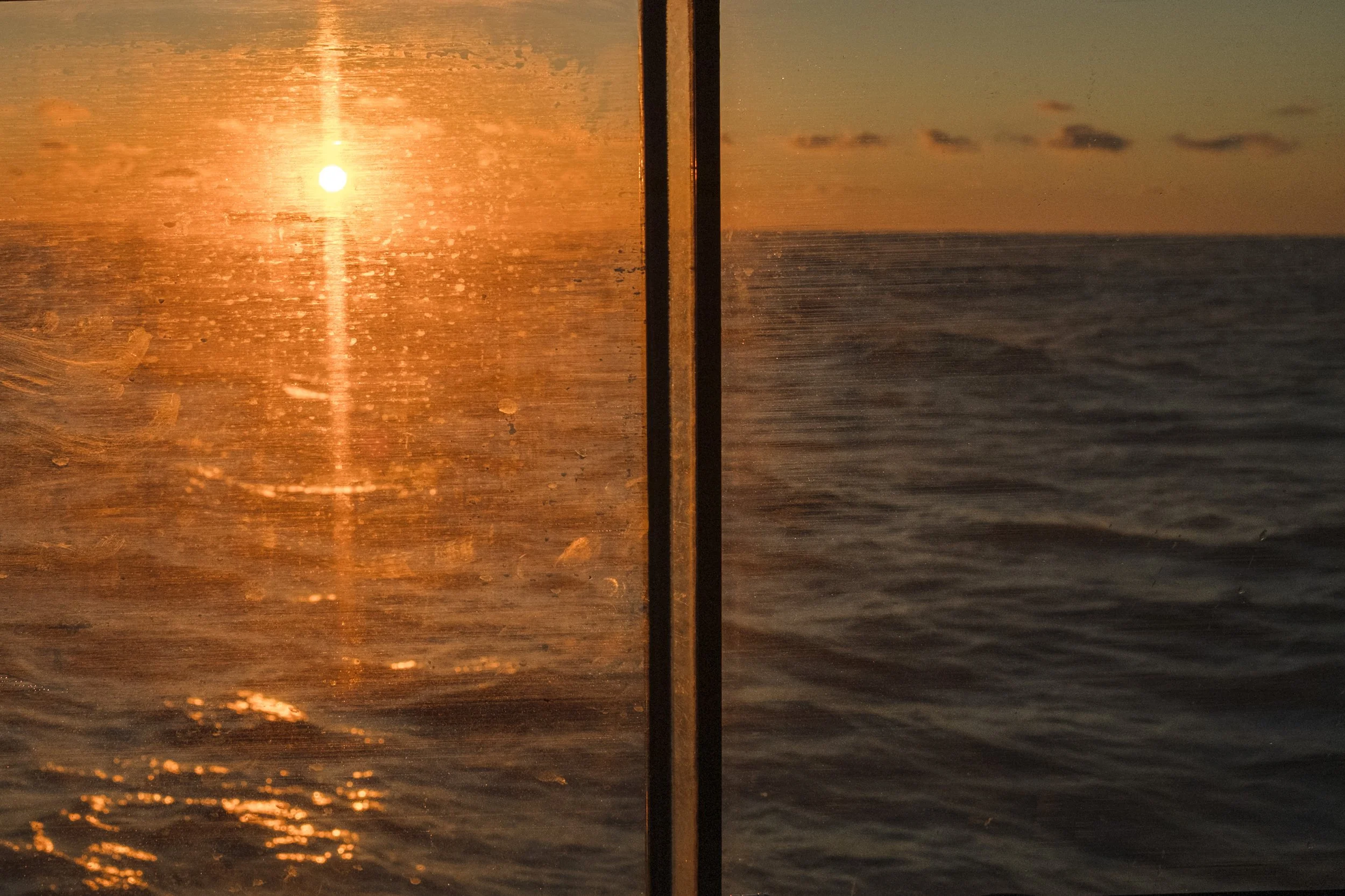 A view of the sea at sunset, seen through a glass window with some water droplets, with a vertical divider in the middle.