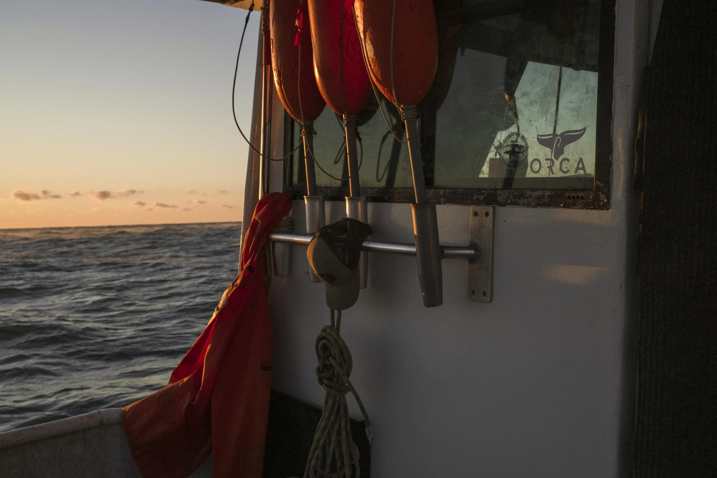 Close-up of a boat's interior with red buoys hanging, a life jacket, and a window with a whale logo that says 'ORCA', against the backdrop of the ocean during sunset.