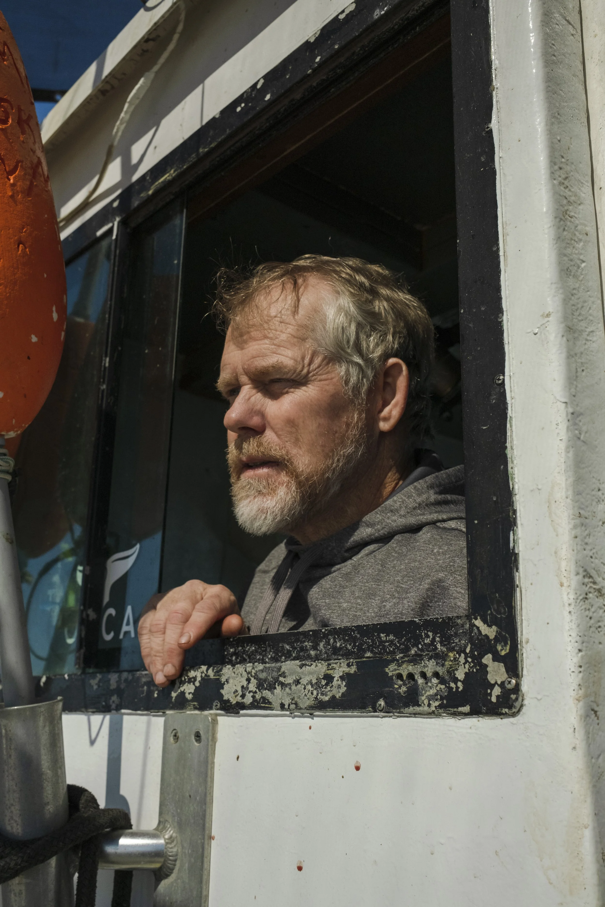 An older man with a beard and gray hair looking out of a small window on a boat or ferry, with a calm expression on his face.