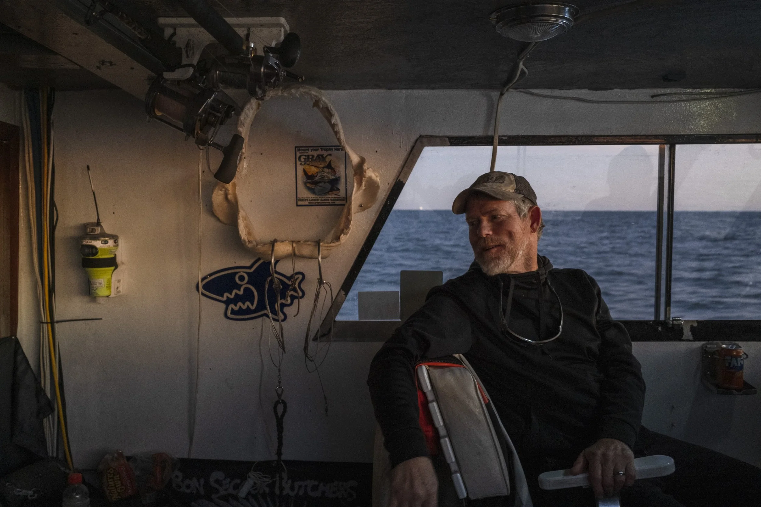 A man with a cap and black jacket sitting inside a boat with the ocean visible through a large window behind him. The boat's interior has fishing gear and a hunting trophy on the wall.