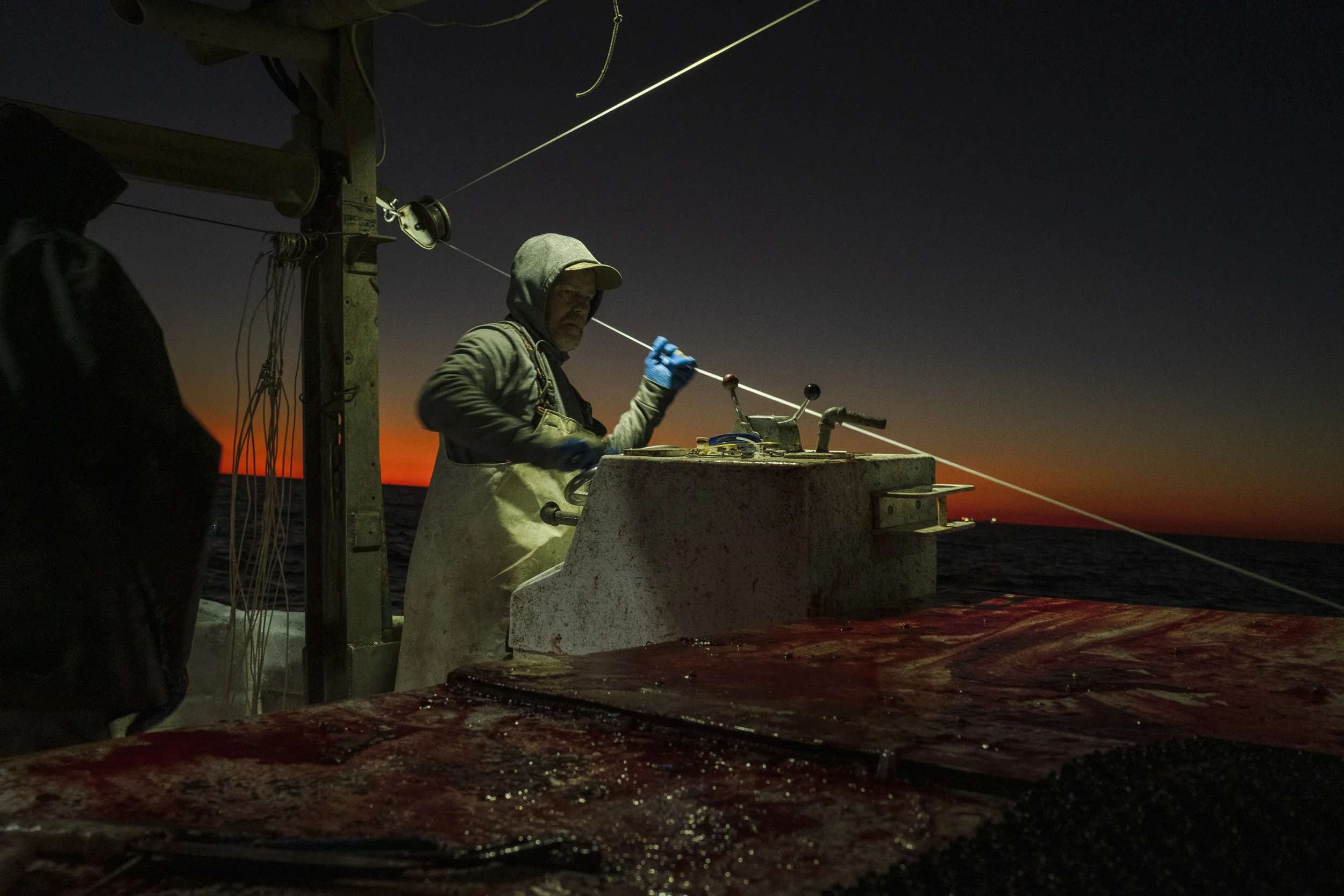 A fisherman on a boat at sunset, wearing a hoodie, apron, and gloves, working with fishing equipment.