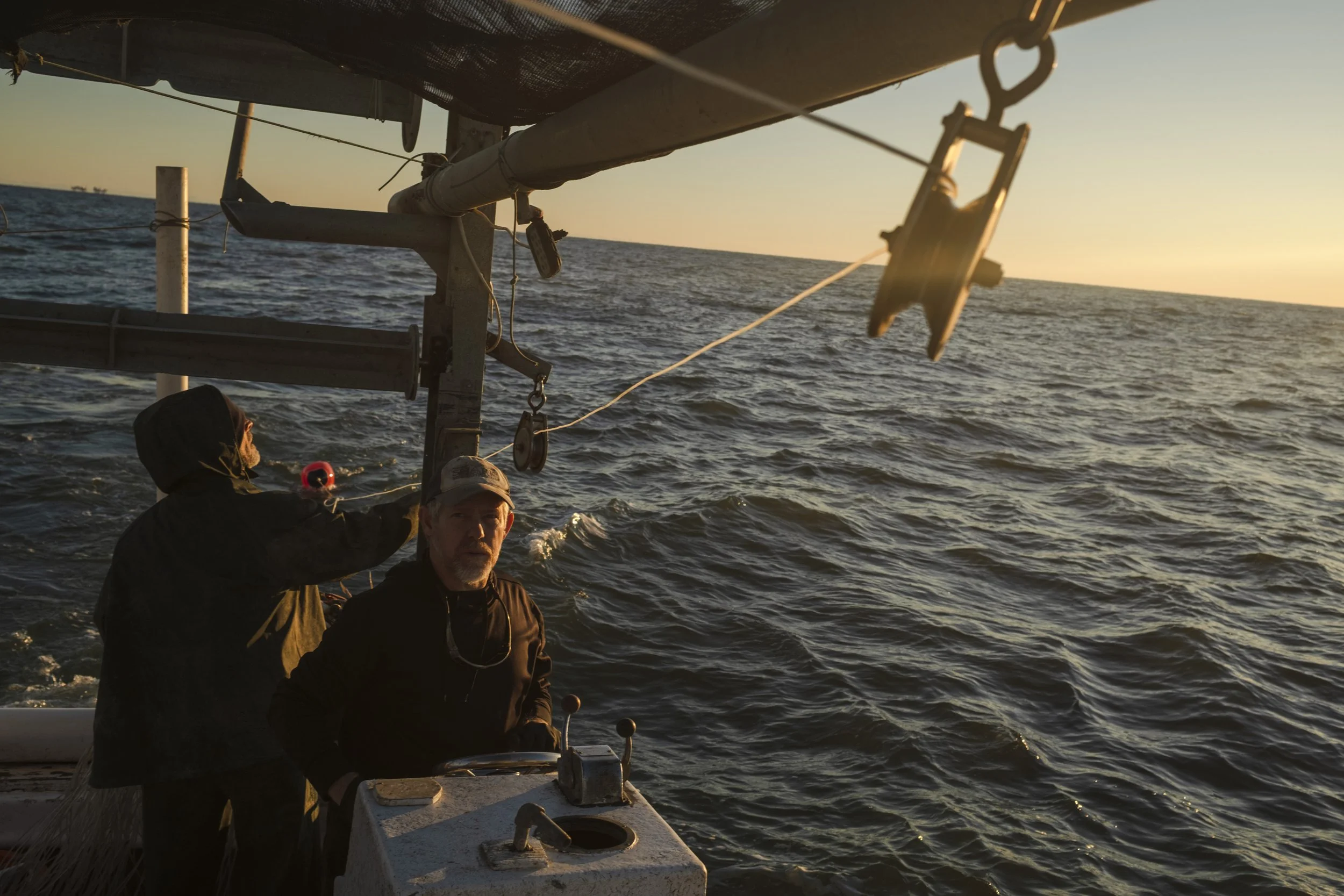Two men on a boat at sunset, one steering and the other working near the mast, with open water and the setting sun in the background.