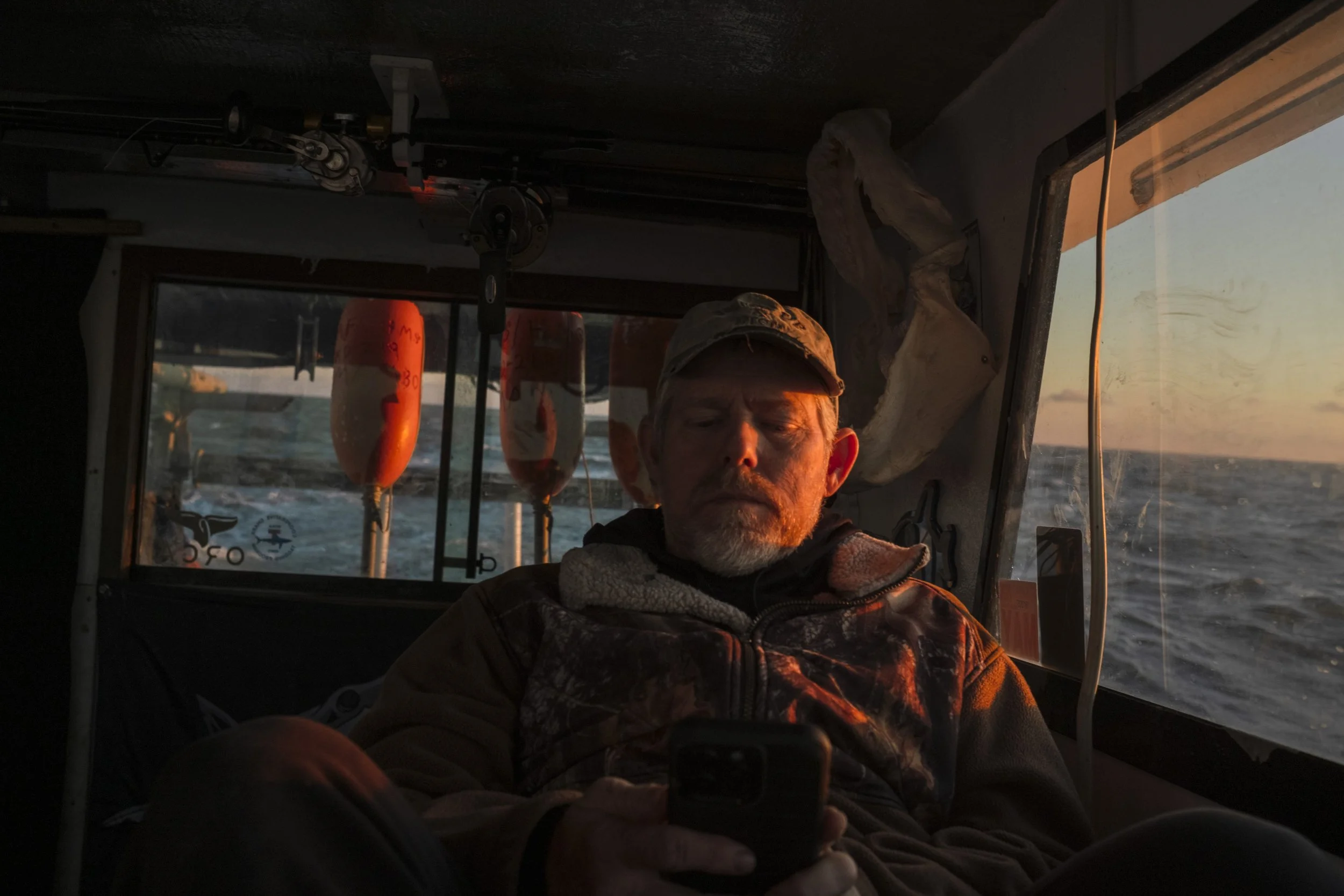 A man with a beard and cap sitting inside a boat, looking at a smartphone, with the ocean visible through the window and the sunset outside.