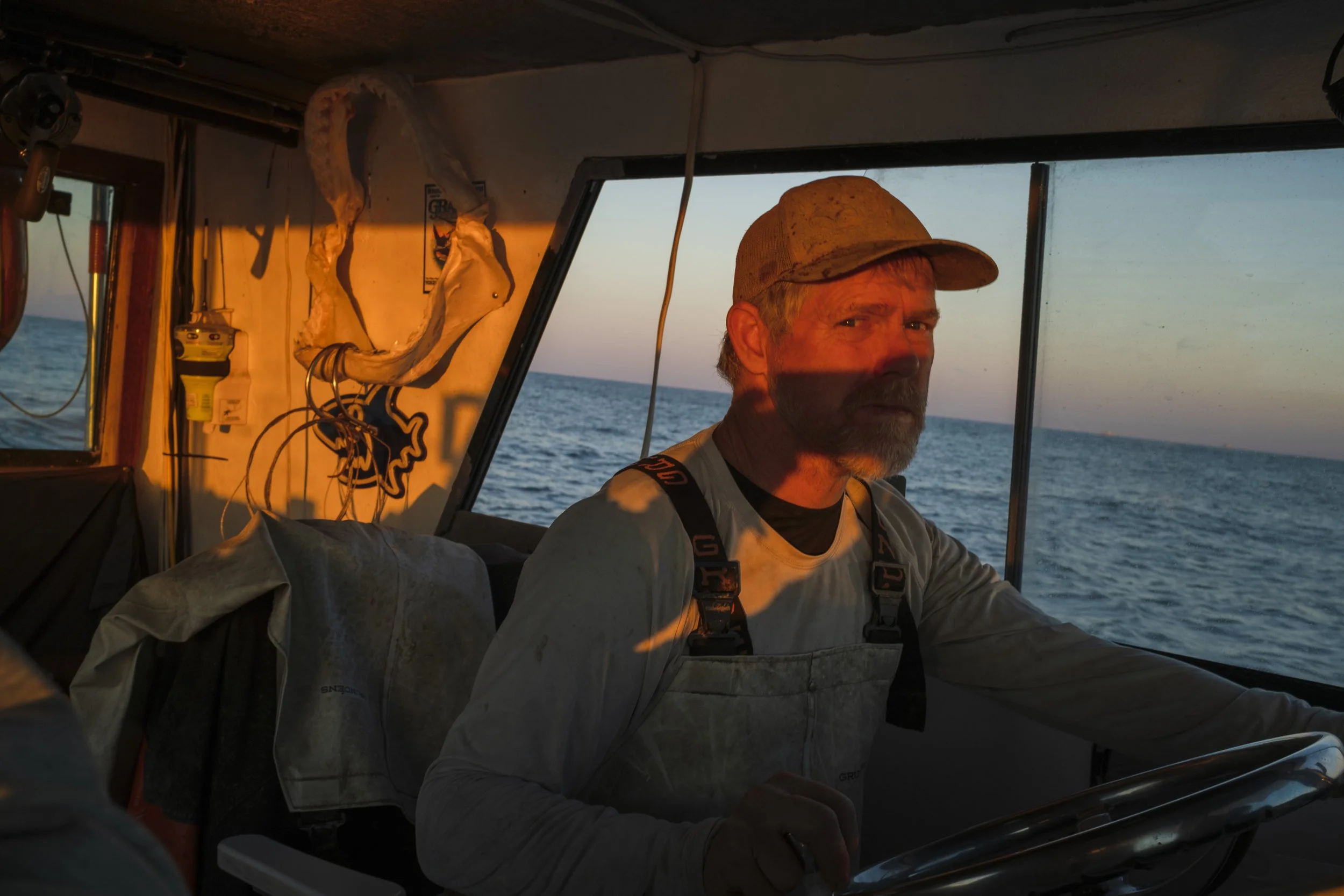 A man with a beard, wearing a baseball cap and overalls, sitting inside a boat during sunset with the ocean in the background.