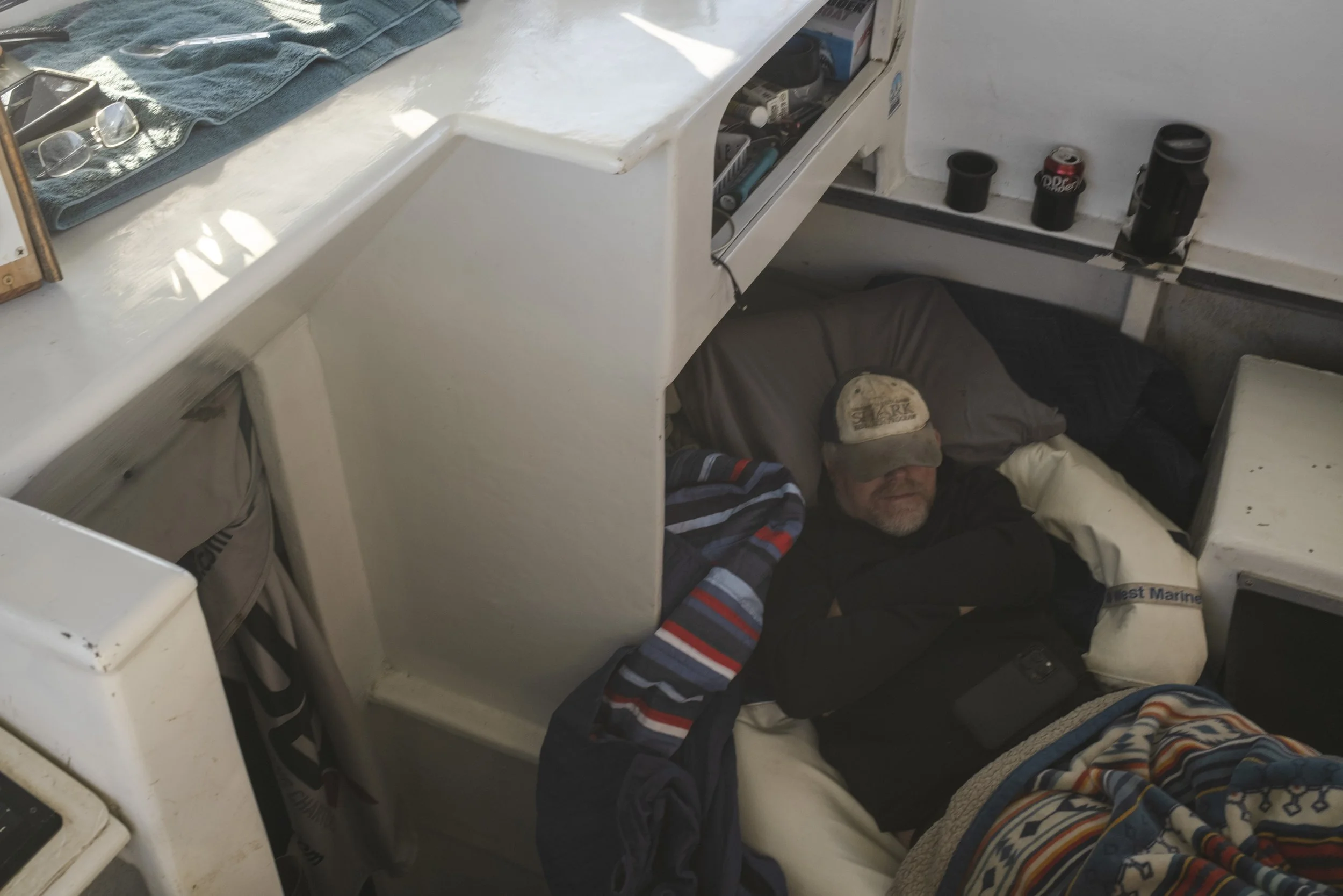 A man sleeping on a bed in a boat, wearing a cap and covered with a blanket, with personal items and beverages nearby.