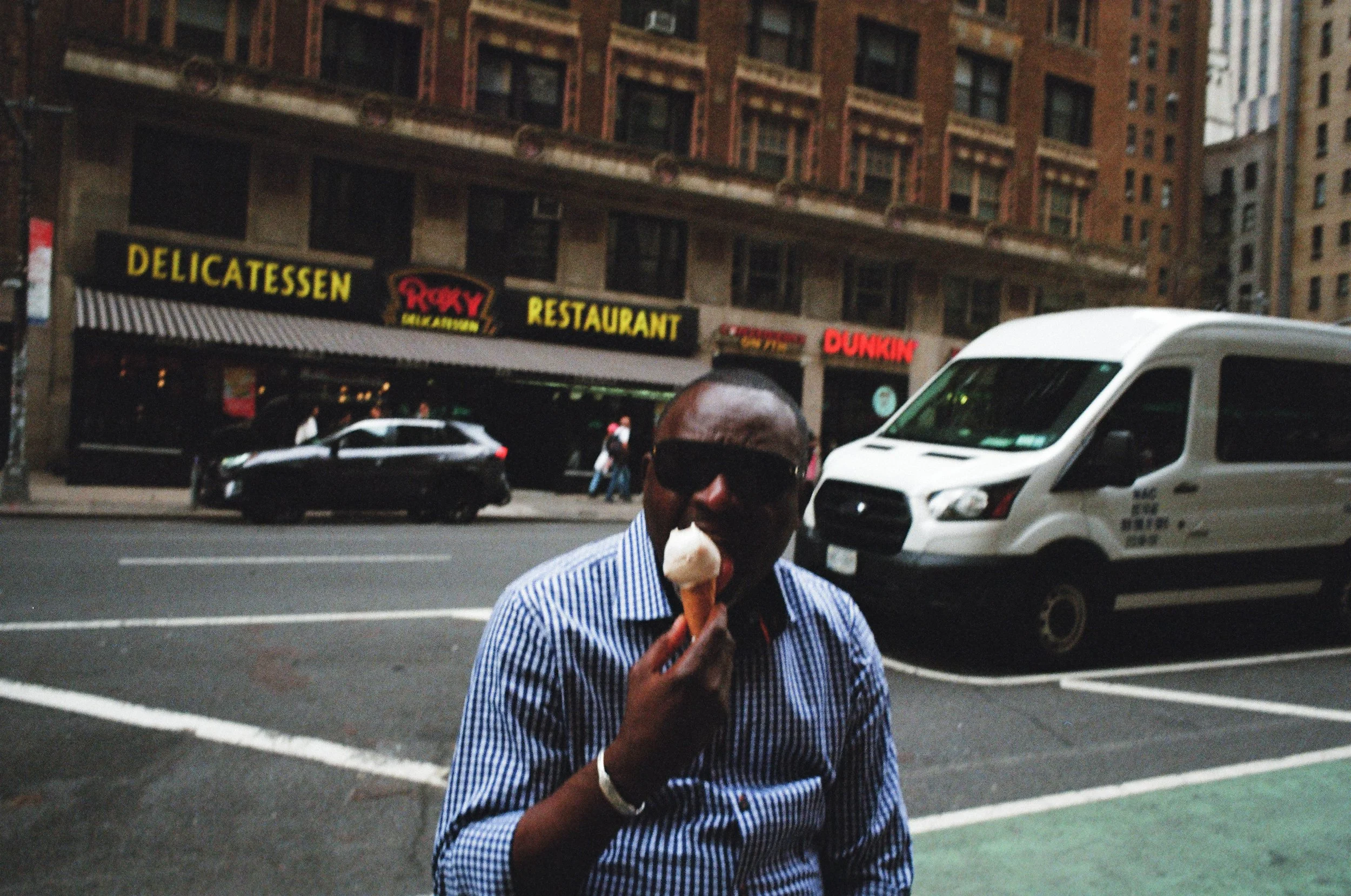 A man wearing sunglasses and a striped shirt eating ice cream on a city street with storefronts for a delicatessen, restaurant, Roxy, Dunkin, and other shops, with cars parked and driving by.