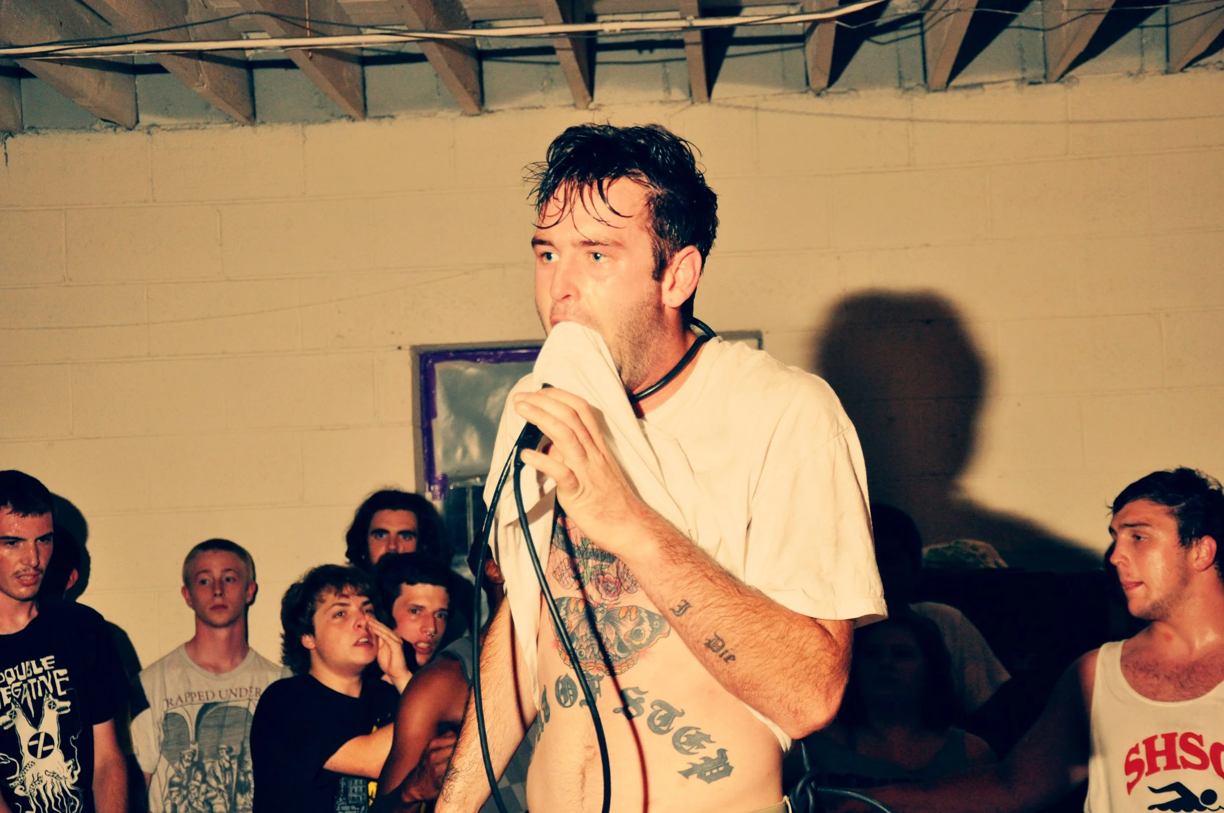 Man with disheveled hair biting a cloth while holding a microphone, surrounded by onlookers in a dimly lit room with a plain wall and a purple-framed mirror in the background.
