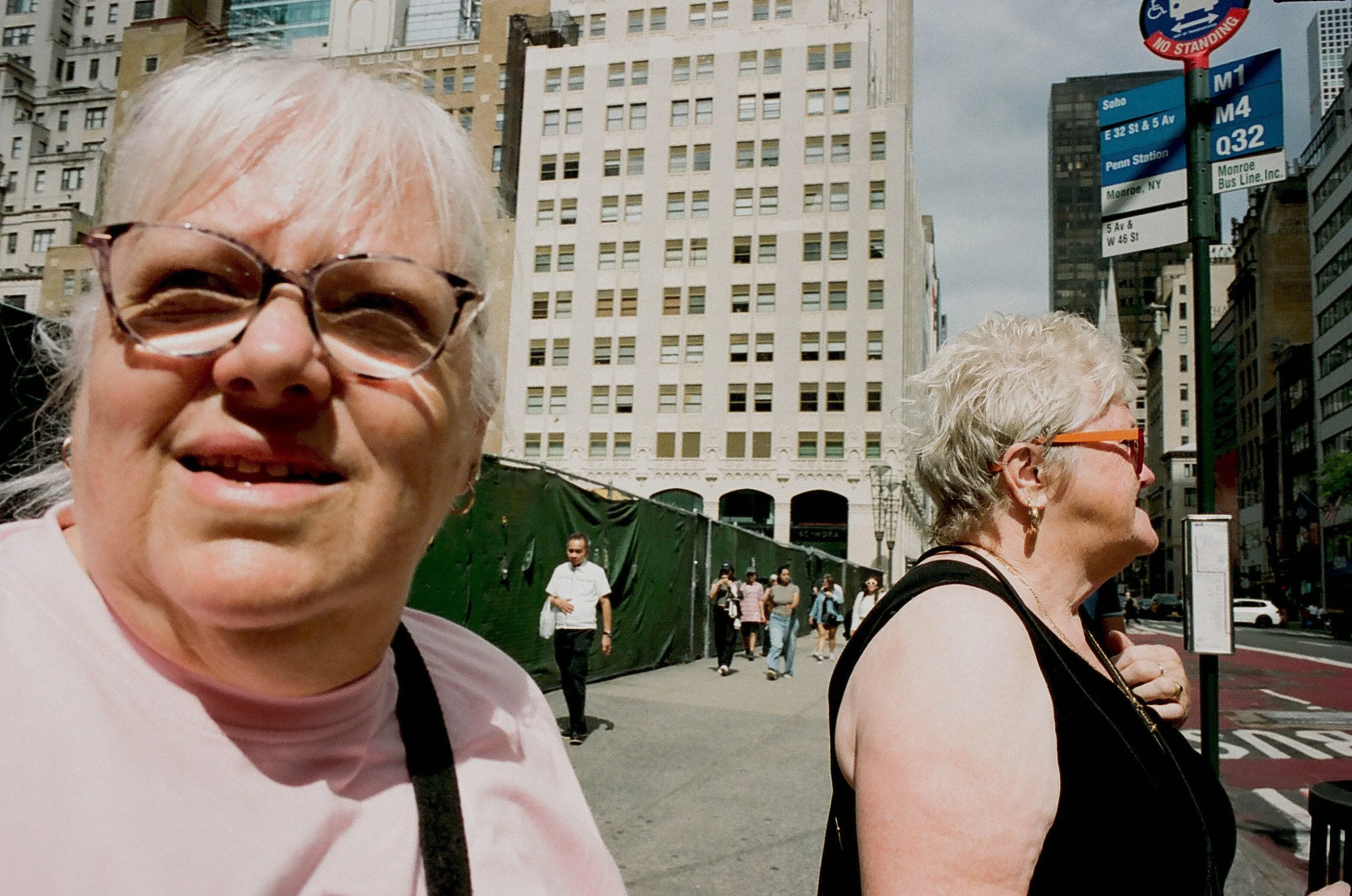 Close-up of two elderly women with short gray hair and glasses walking on a busy city street with tall buildings and signs, and other pedestrians in the background.