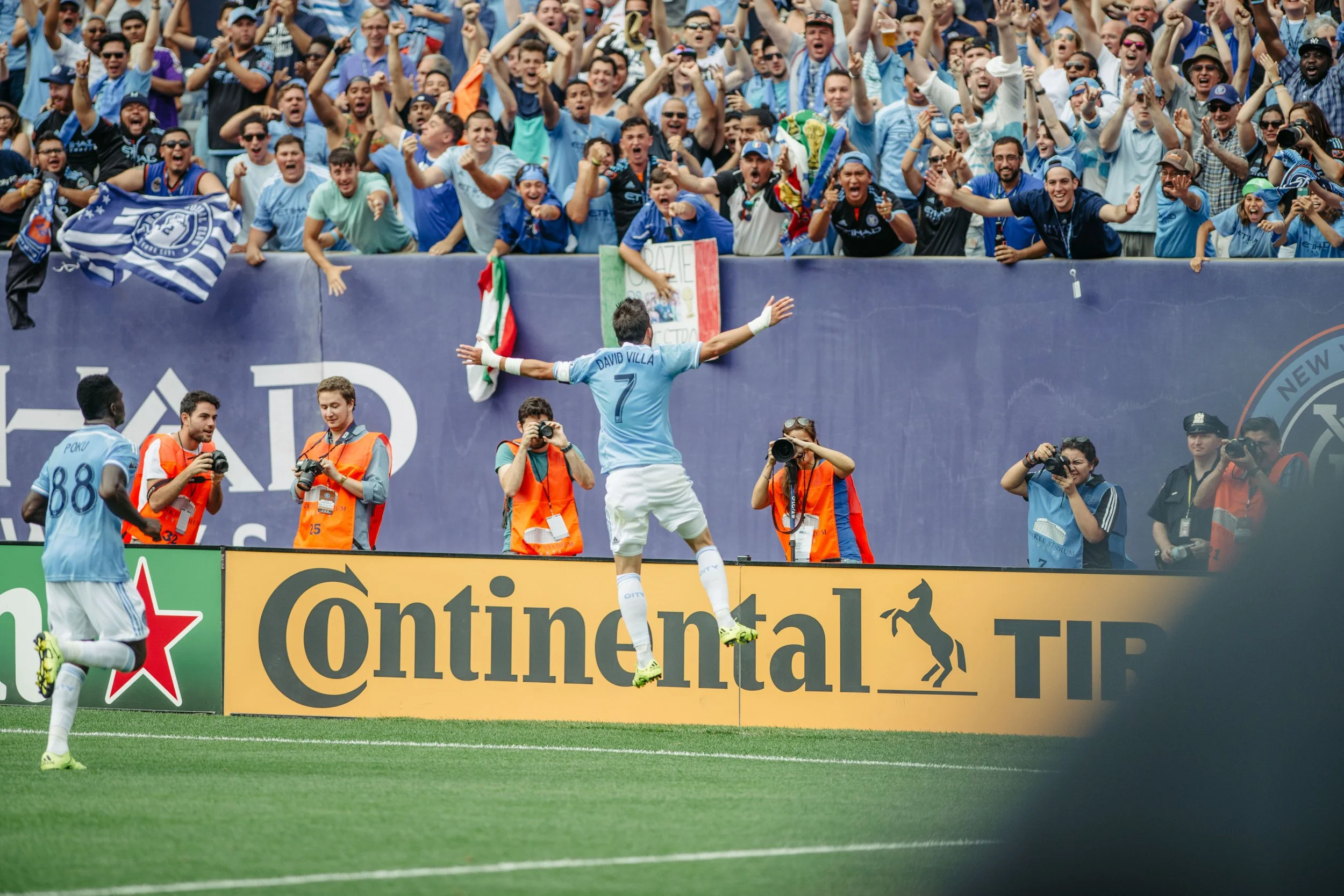Soccer player David Villa celebrating by jumping near the sideline in front of cheering fans, with photographers capturing the moment.