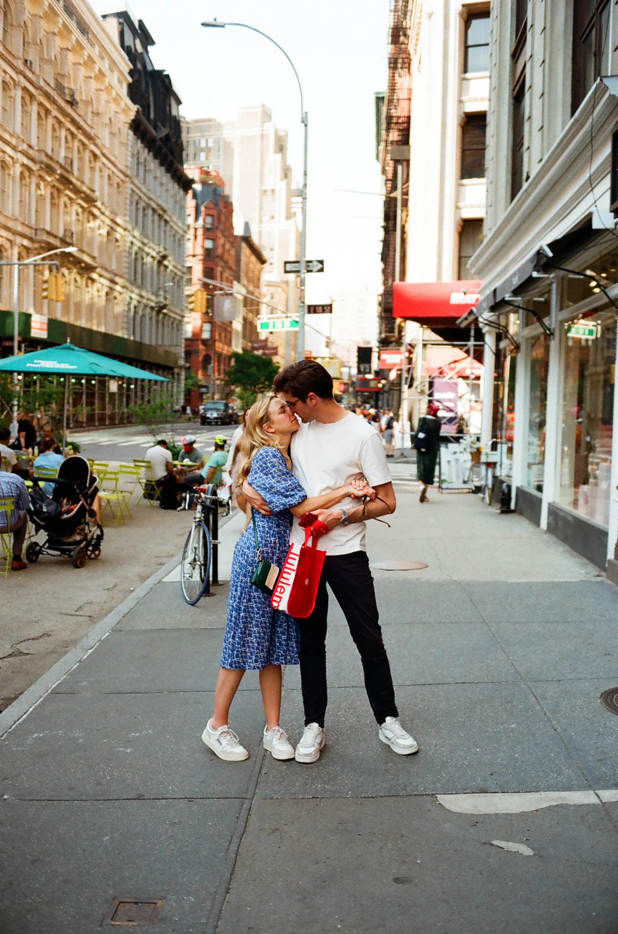 A young couple sharing a kiss on a city sidewalk during daylight, with a woman in a blue dress and a man in a white t-shirt and black pants, surrounded by buildings, street signs, and outdoor seating.