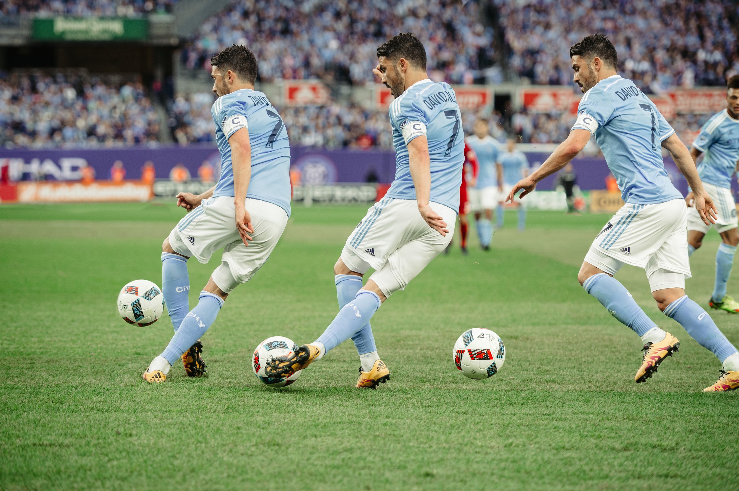 Three soccer players in light blue jerseys and white shorts practice passing with soccer balls on a green field, with a crowd watching in the background.