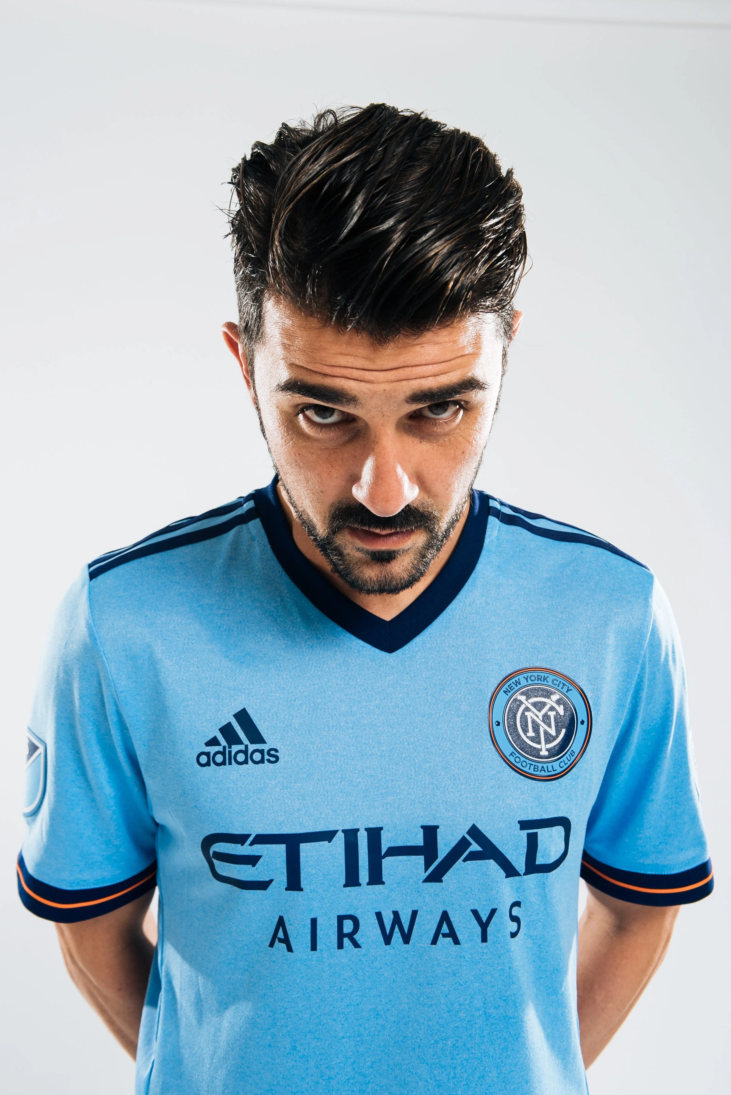 A man with dark hair and beard wearing a light blue NYC football club jersey, looking upward at the camera with a serious expression.