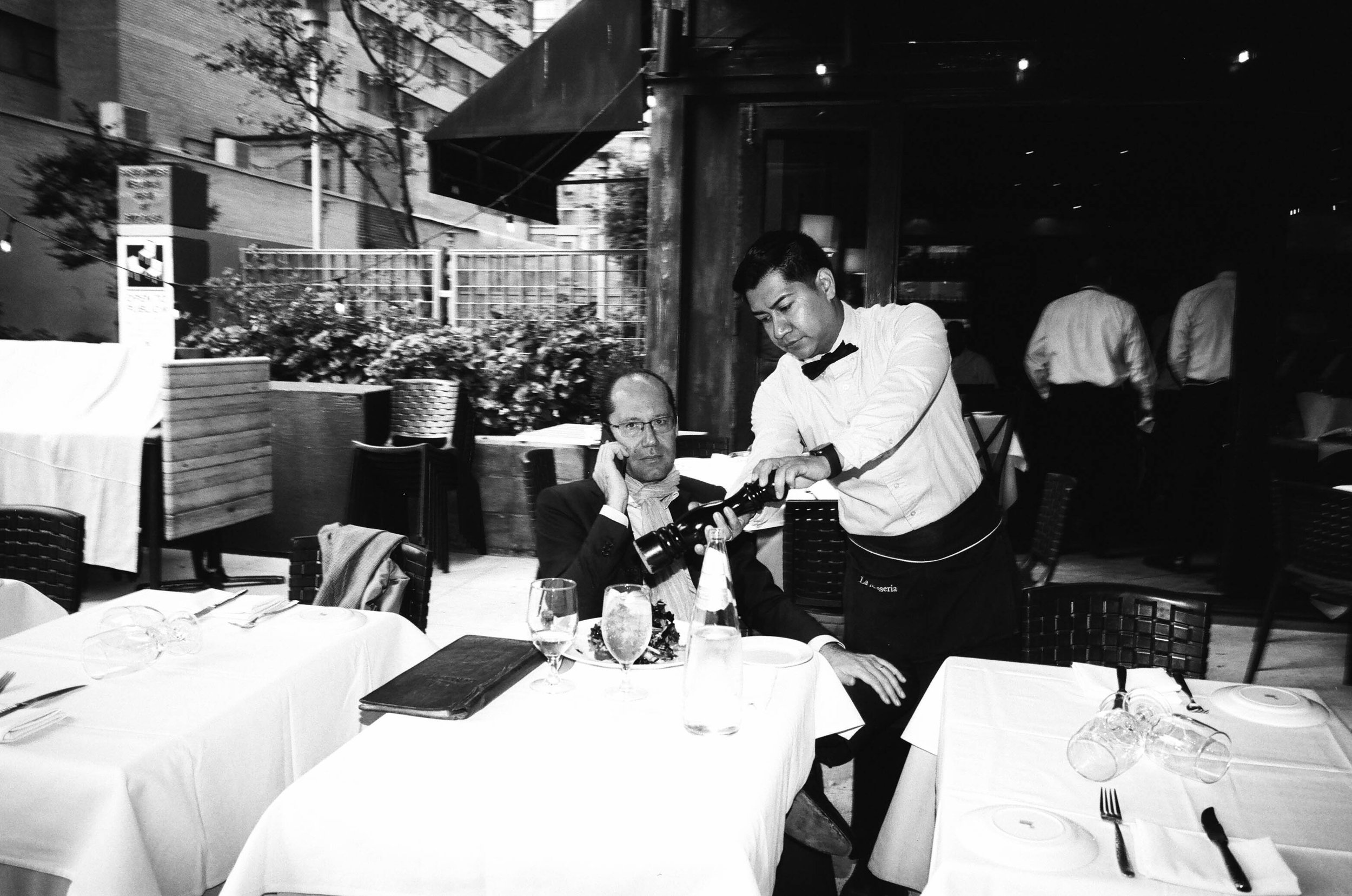 A waiter clearing an empty table at a restaurant as a man in glasses and a suit sits at the table, holding a glass of water, with a fancy dish in front of him. There are additional tables, chairs, and other waiters in the background.