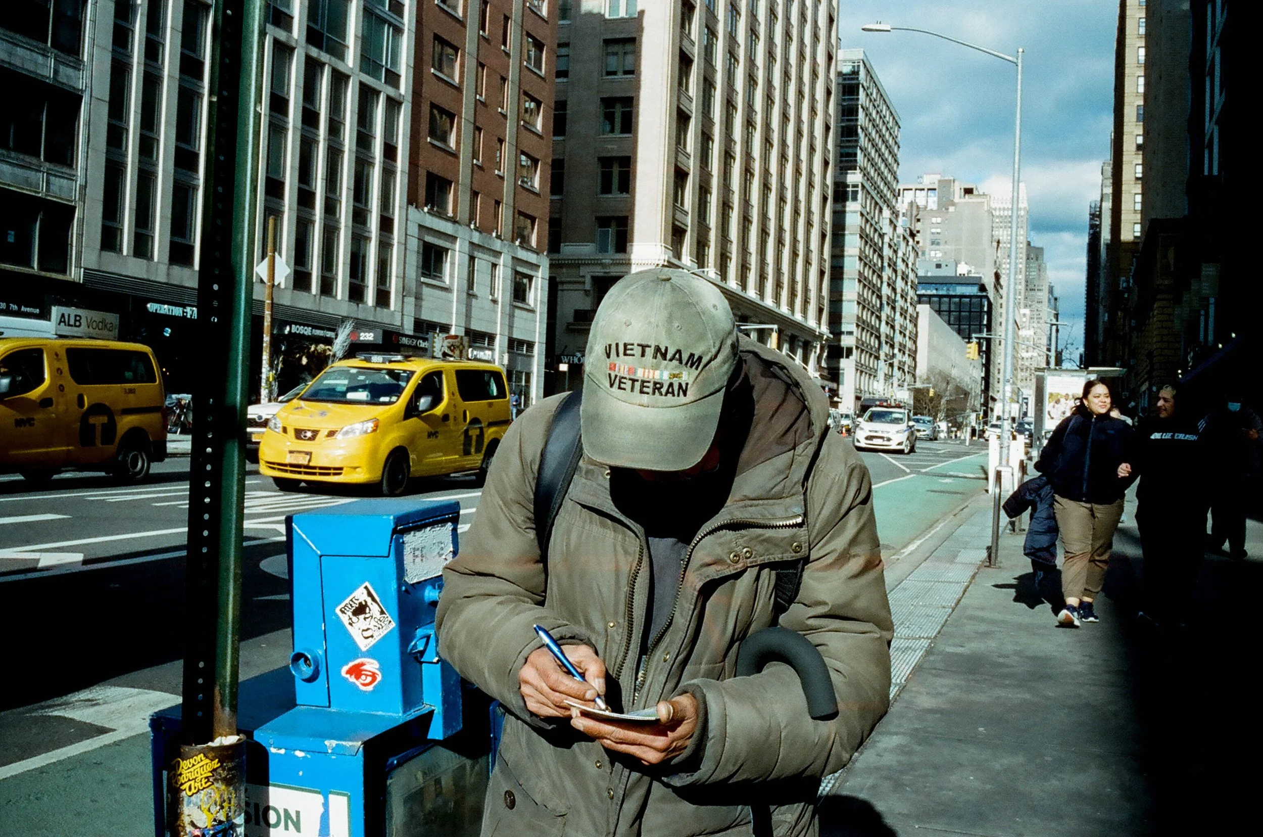 A man wearing a tan jacket and a beige cap with 'Vietnam Veteran' embroidered on it is writing on a notepad while holding a cane on a busy city sidewalk with yellow taxis and tall buildings.