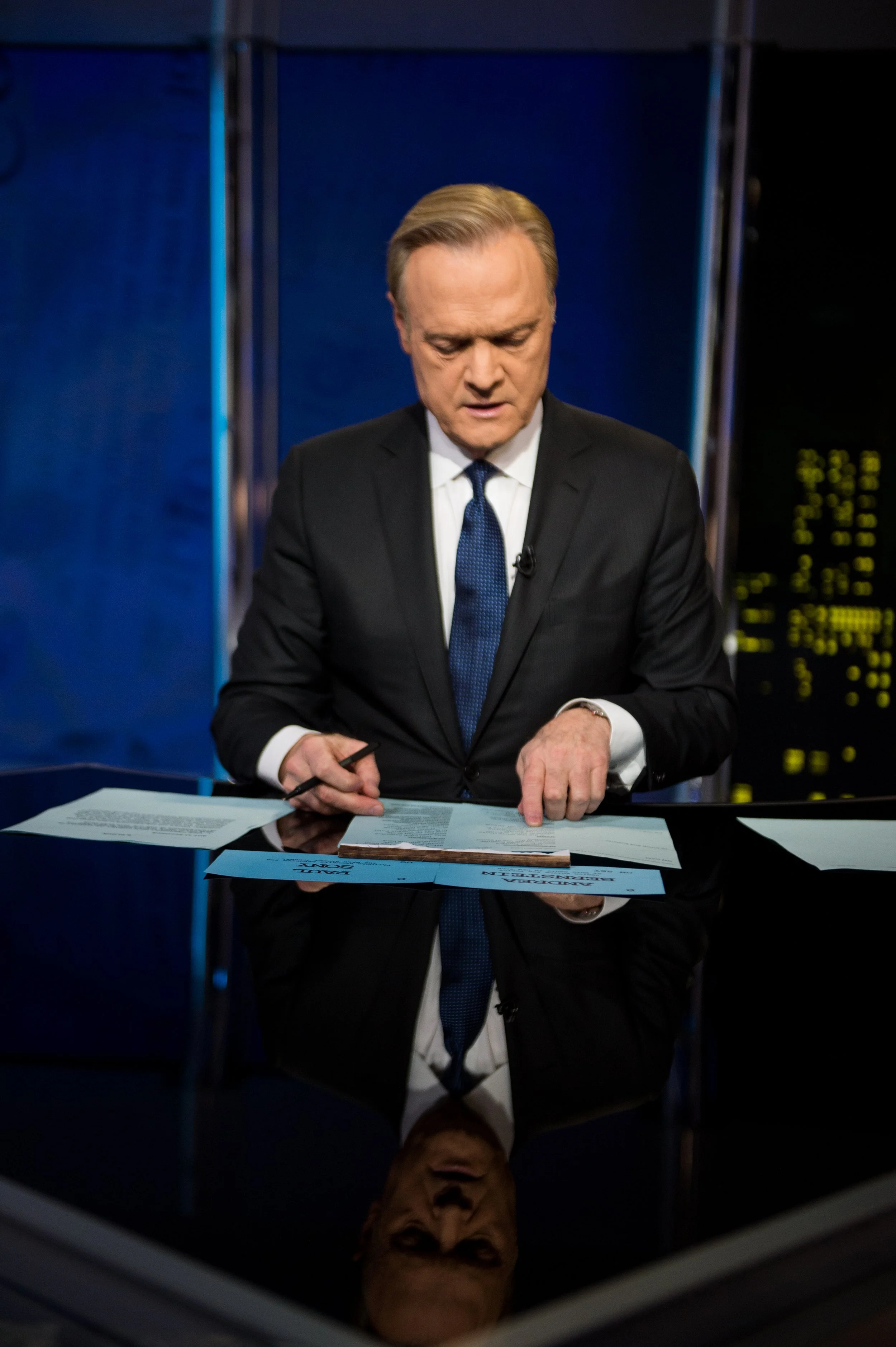 A man in a black suit and blue tie sitting at a glossy black table, reading documents, with a cityscape illuminated at night in the background.