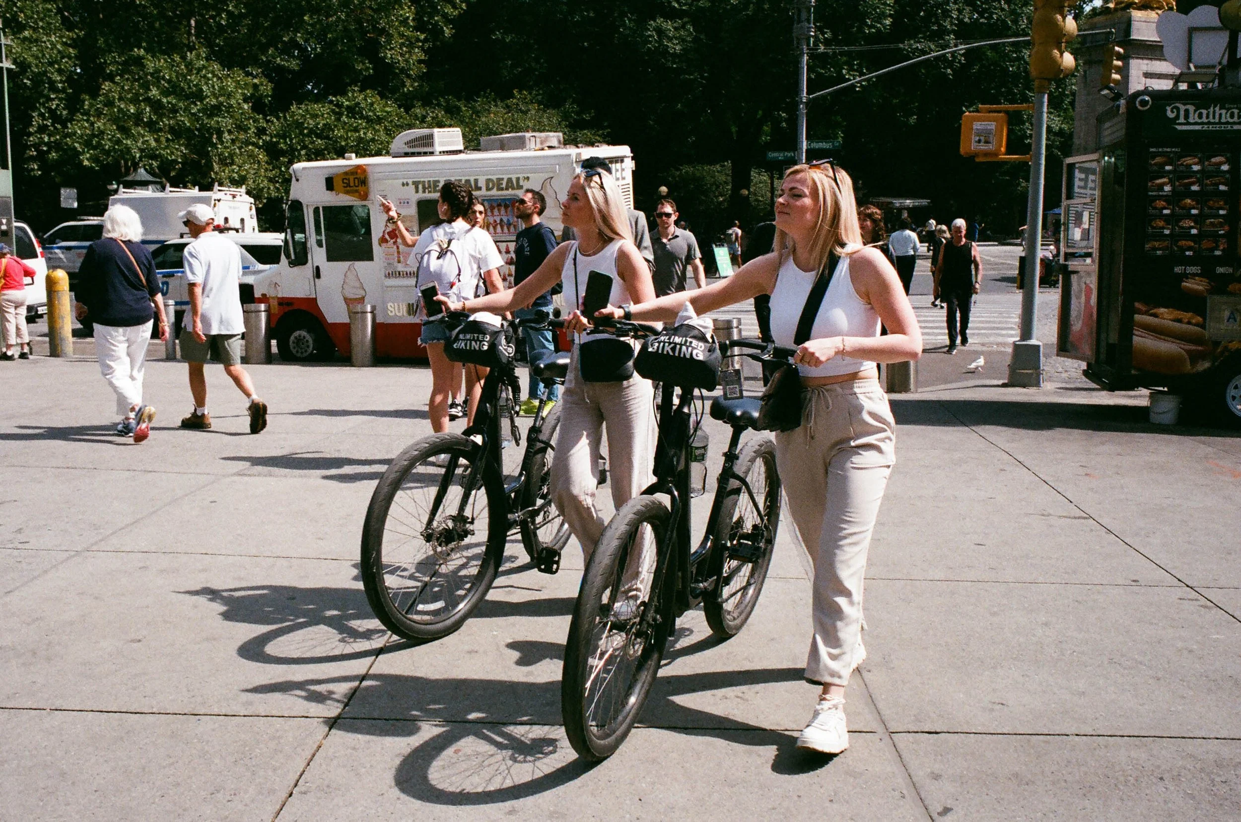 Two women in white tank tops and beige pants walking with black bikes on a city sidewalk.