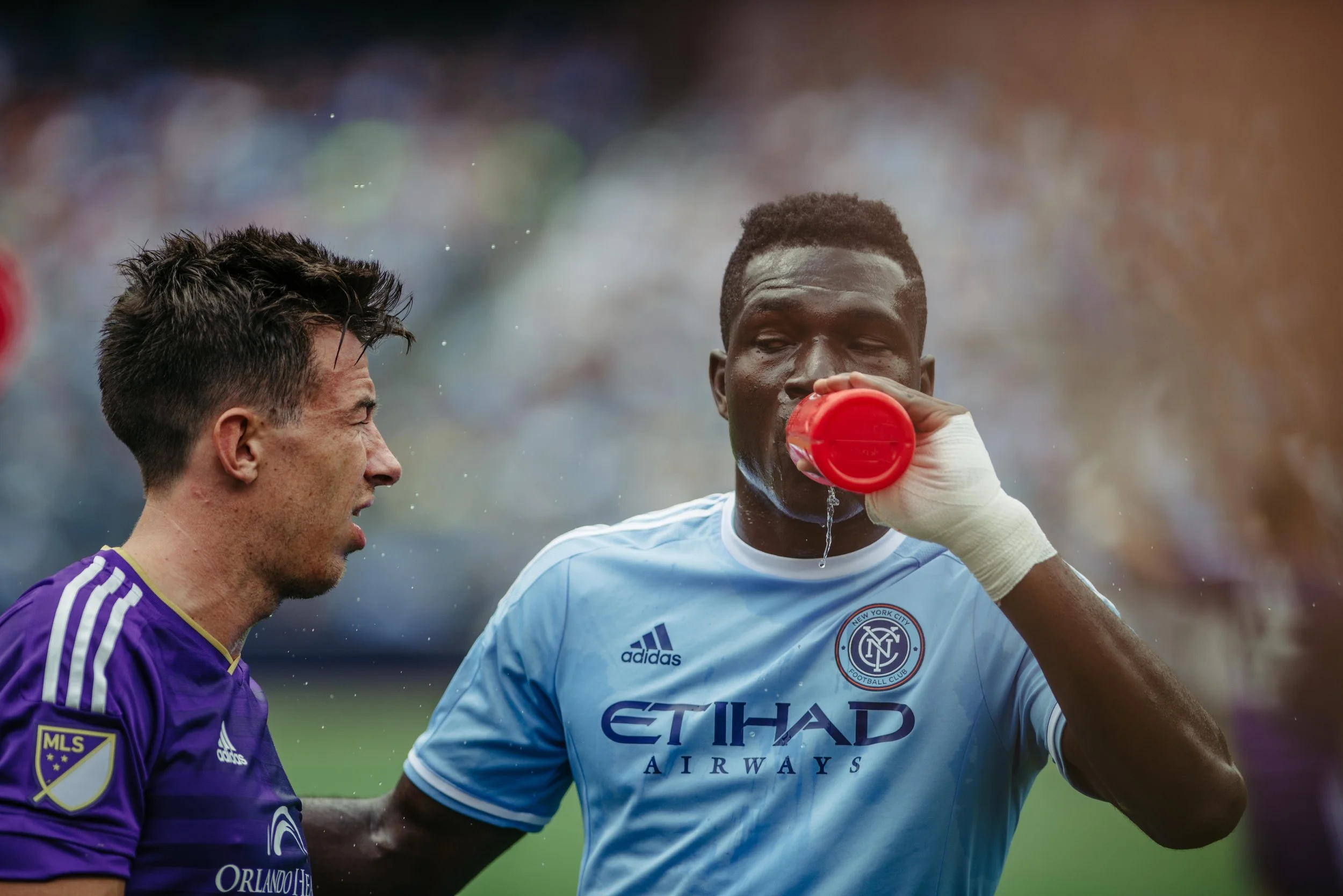 Two soccer players on the field, one in purple jersey and the other in light blue jersey, hydrating with water bottles during a game.