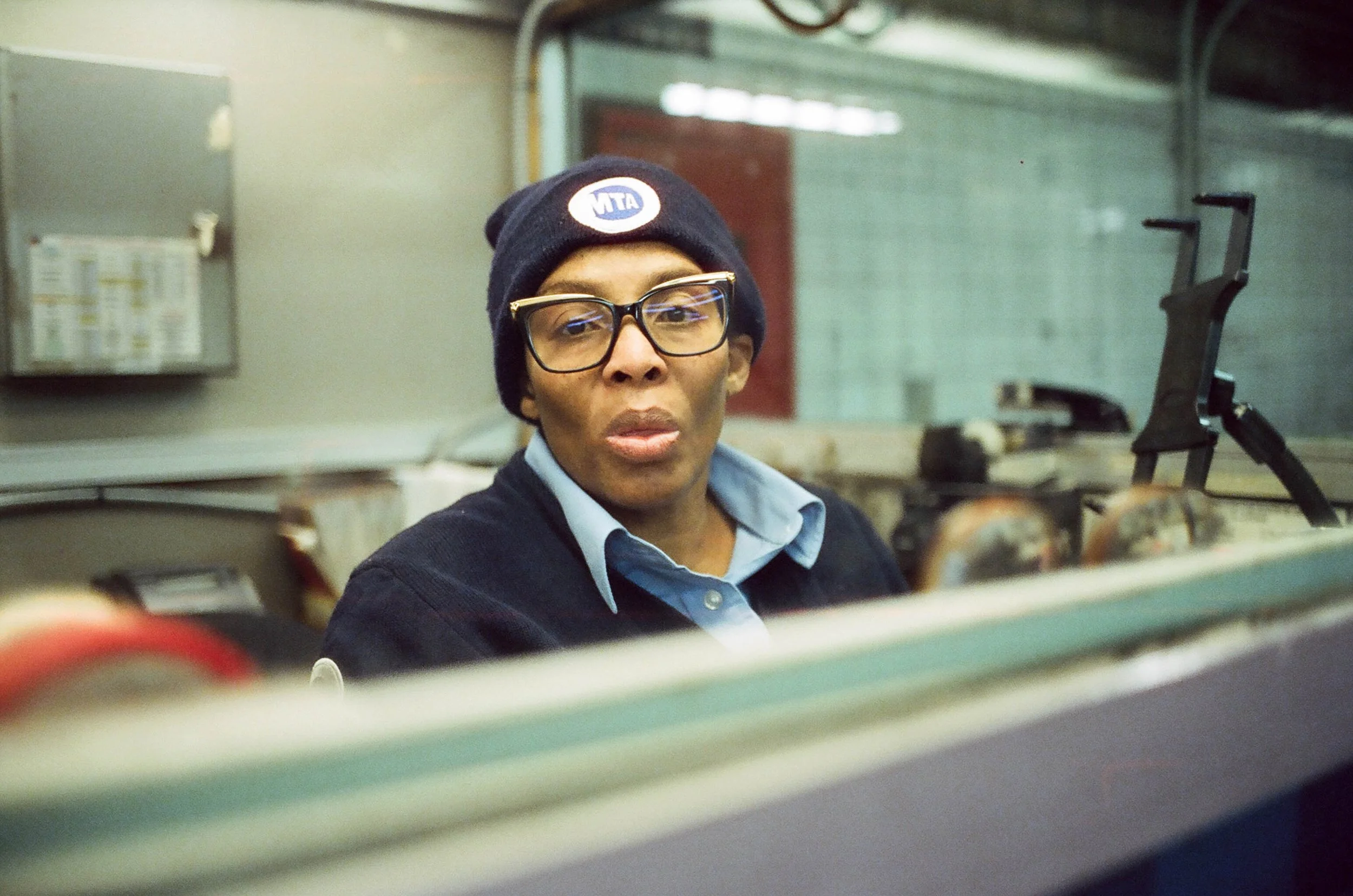 A person wearing glasses, a navy beanie with an 'MTA' logo, a light blue collared shirt, and a black sweater, appears to be working or observing from behind a counter or window, with subway tunnels in the background.