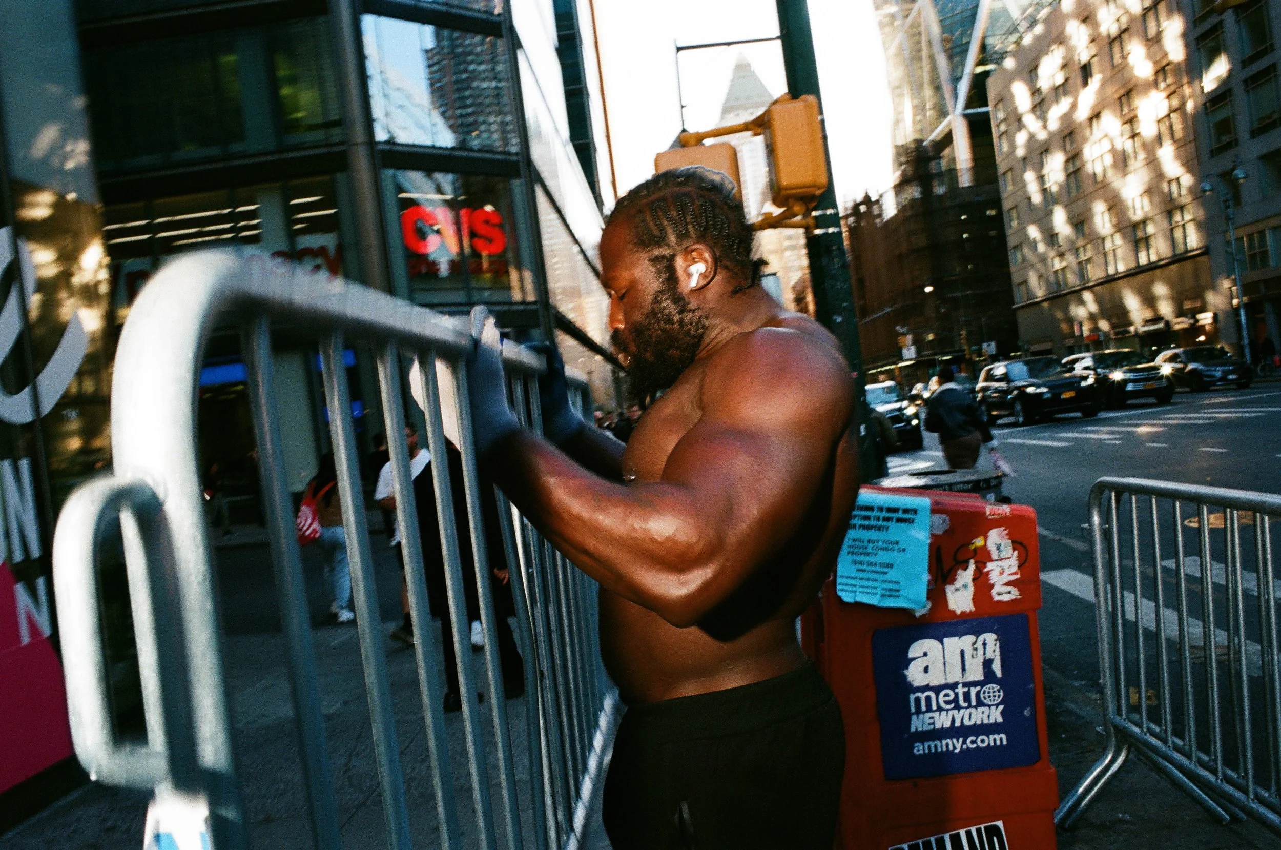 A shirtless man with a beard and short hair, wearing white earbuds, adjusting a metal barricade on a busy city street, with tall buildings and traffic in the background.
