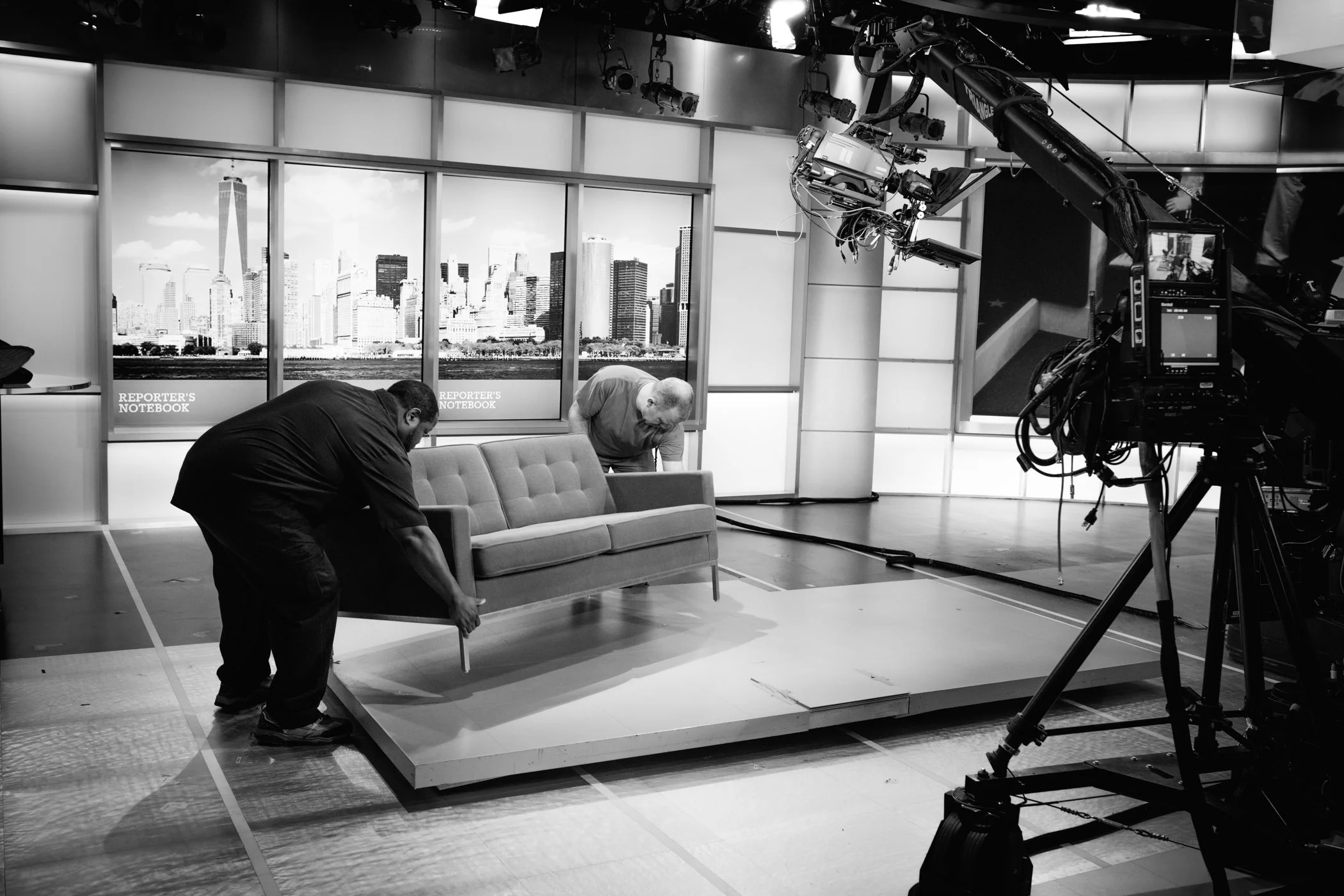 Two men setting up a television studio with a backdrop of a city skyline, a couch on a platform, and professional filming equipment.