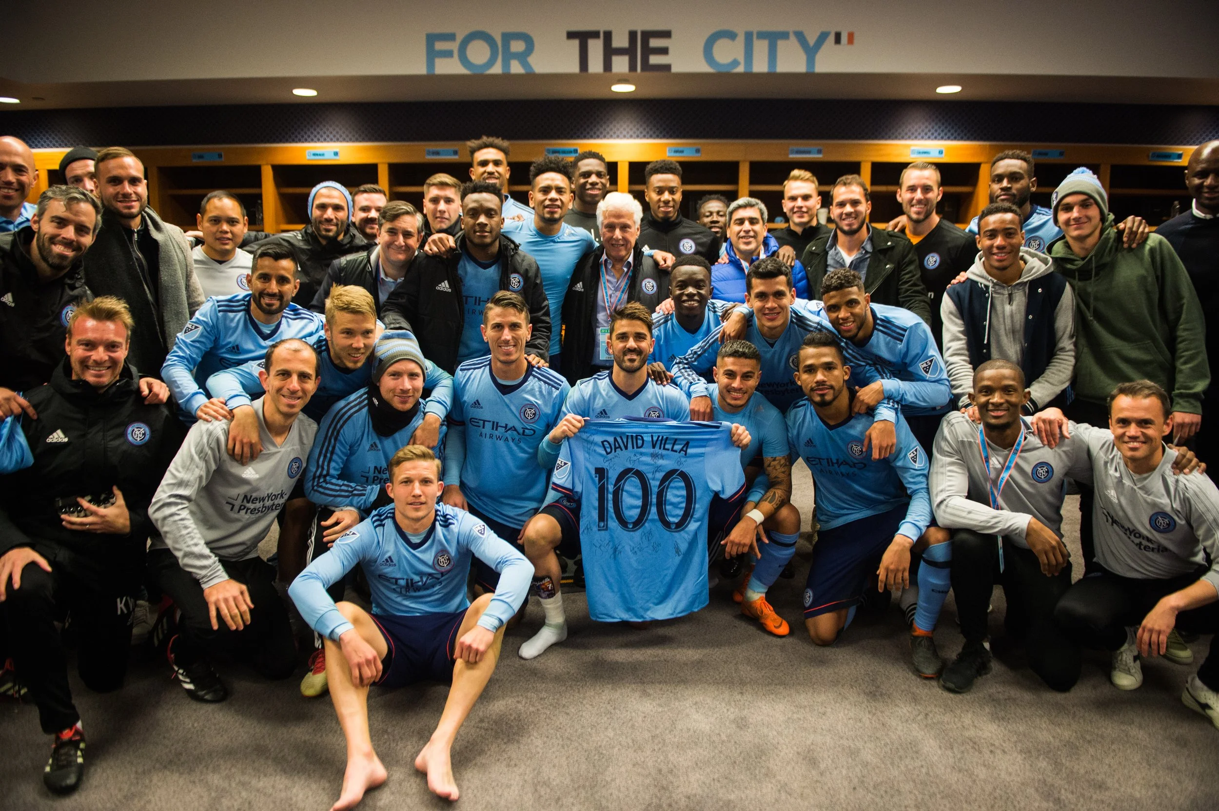 Group of soccer players and staff in locker room, celebrating successful game. One player holds a jersey with "David Villa 100" written on it.