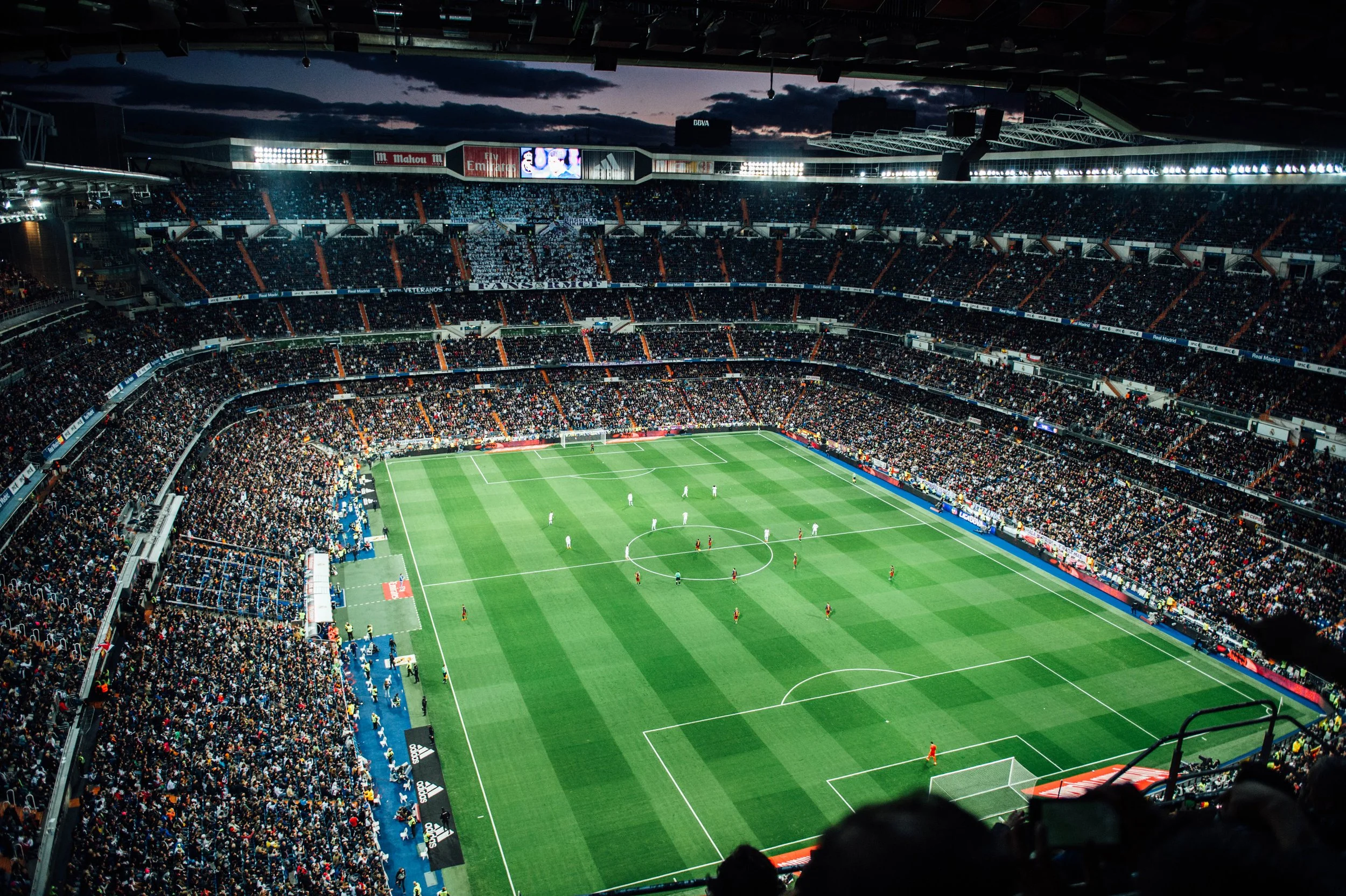 A crowded soccer stadium filled with spectators watching a game at dusk, with players on the field.