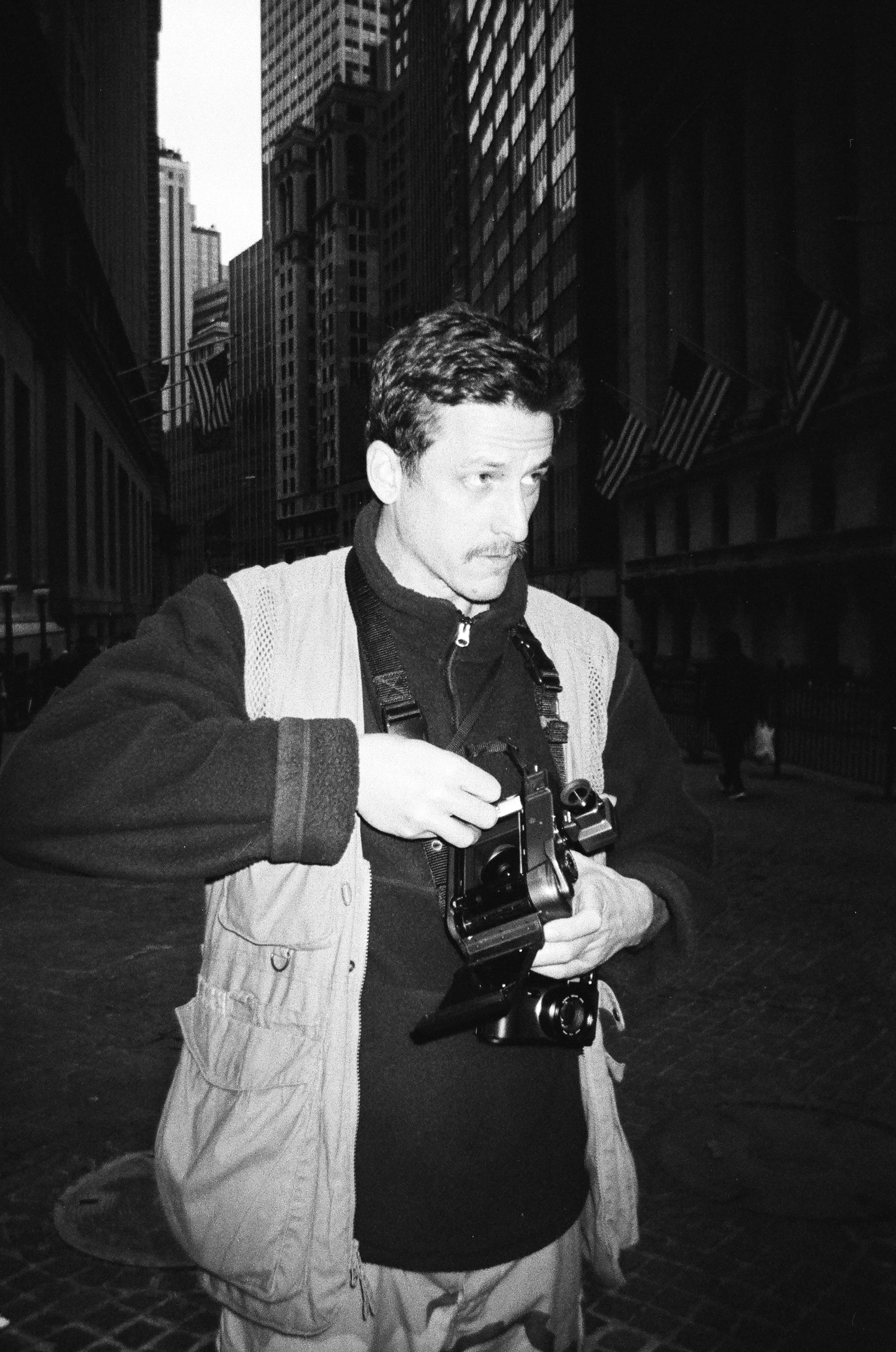 Man holding a camera on a city street with tall buildings and American flags in the background.