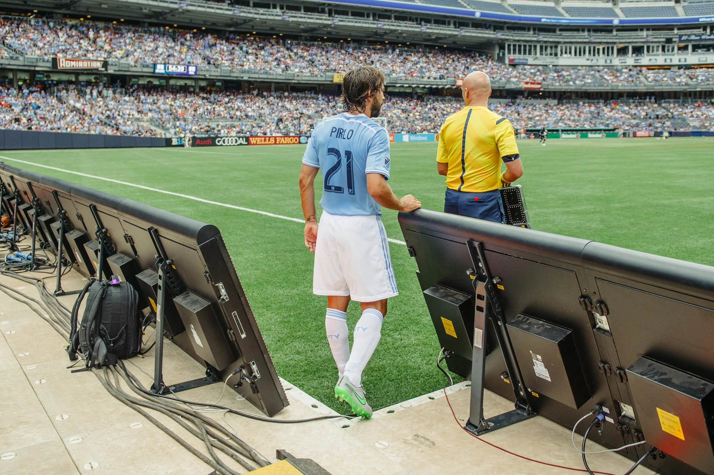 Soccer player in light blue uniform walking near sideline with video assistant referee, in a crowded stadium.