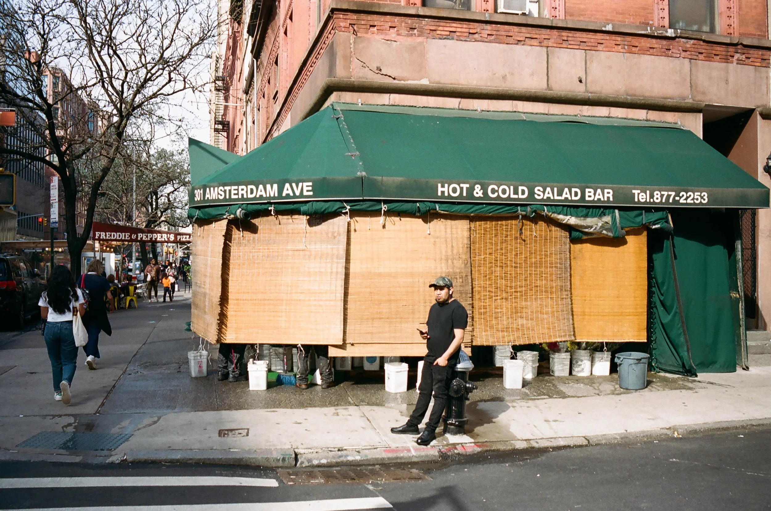 Street corner with a closed food stand under a green awning that reads '301 Amsterdam Ave Hot & Cold Salad Bar Tel.877-2253'. Man in black clothing and cap standing near the stand, looking at phone. Pedestrians walking by on sidewalk, storefronts and