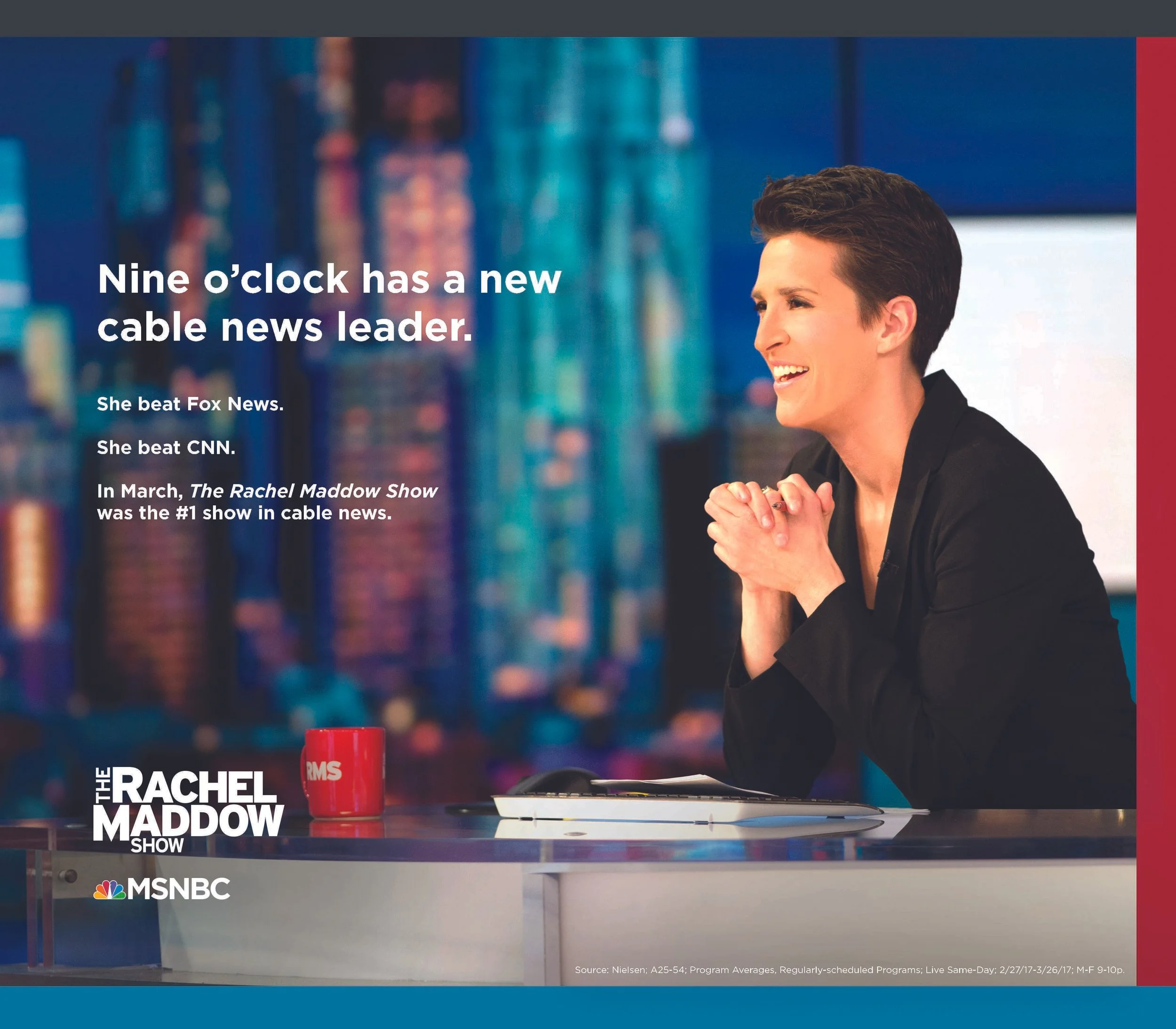 A woman with short hair smiling on the set of 'The Rachel Maddow Show' at MSNBC. The background shows colorful blurred lights, and there is a red mug with white letters on the desk in front of her.