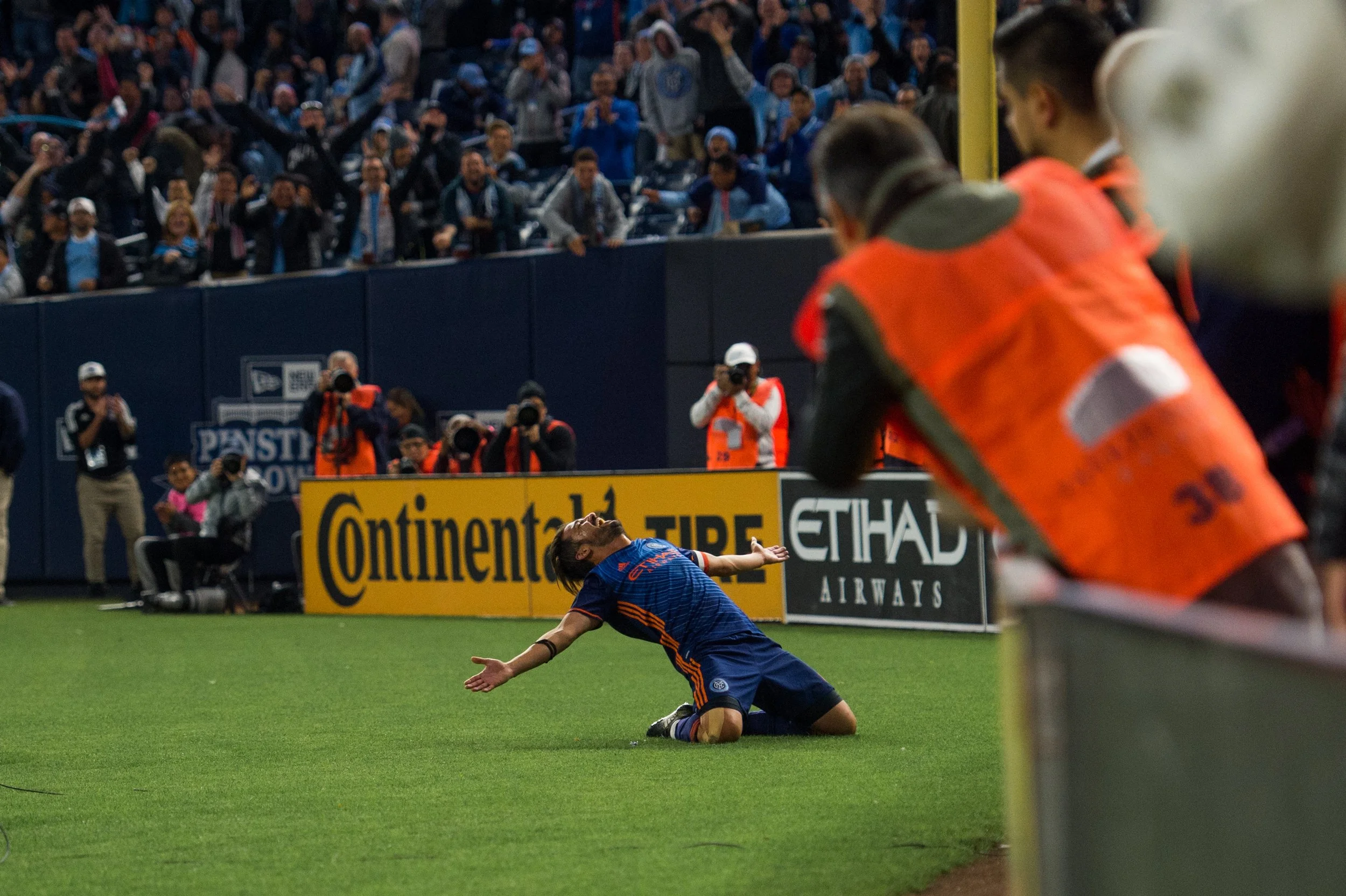 A soccer player is kneeling on the field with arms outstretched, celebrating in front of a crowd at a stadium, while photographers and staff capture the moment.