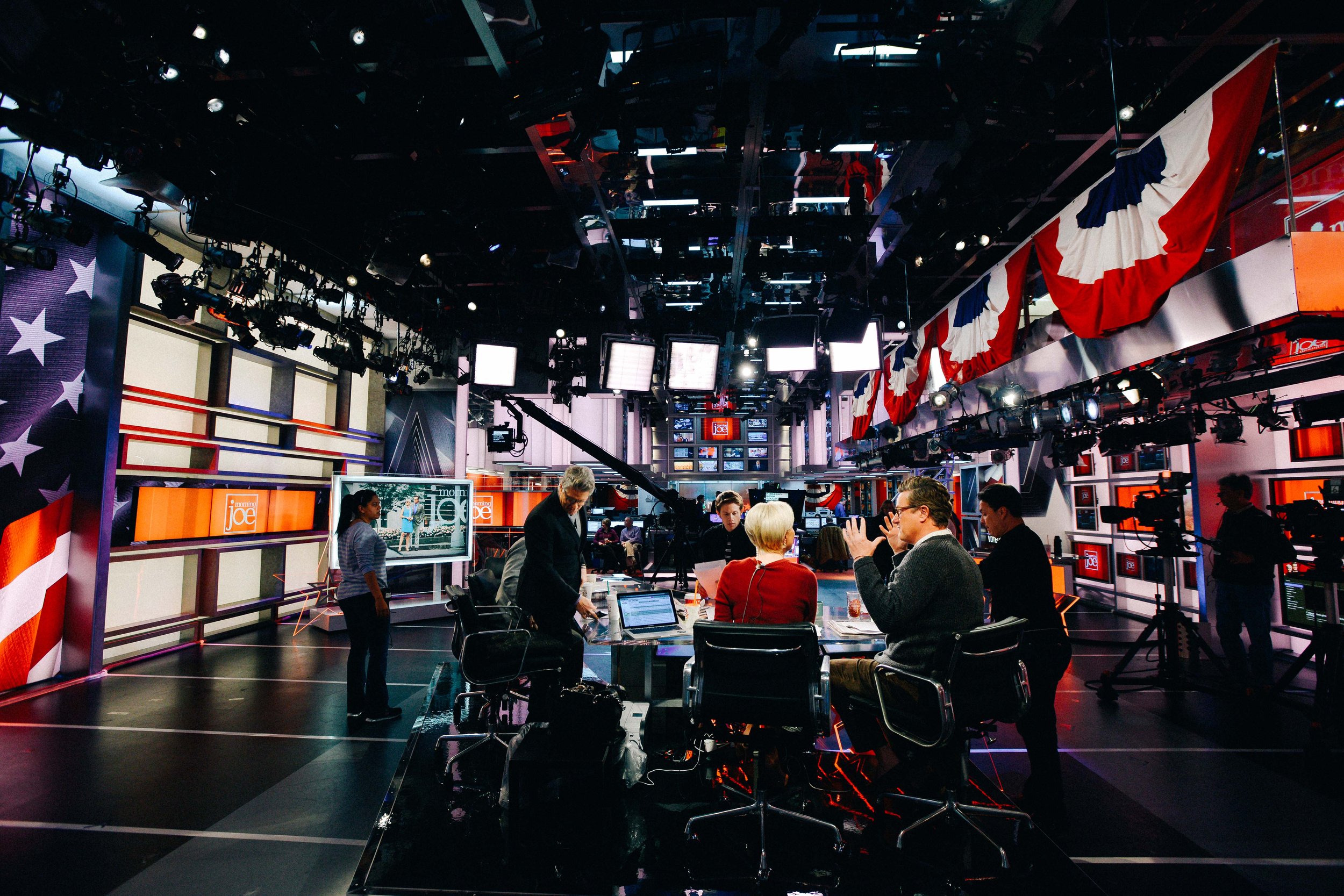 TV news studio with crew members, cameras, and large screens, decorated with patriotic red, white, and blue bunting.